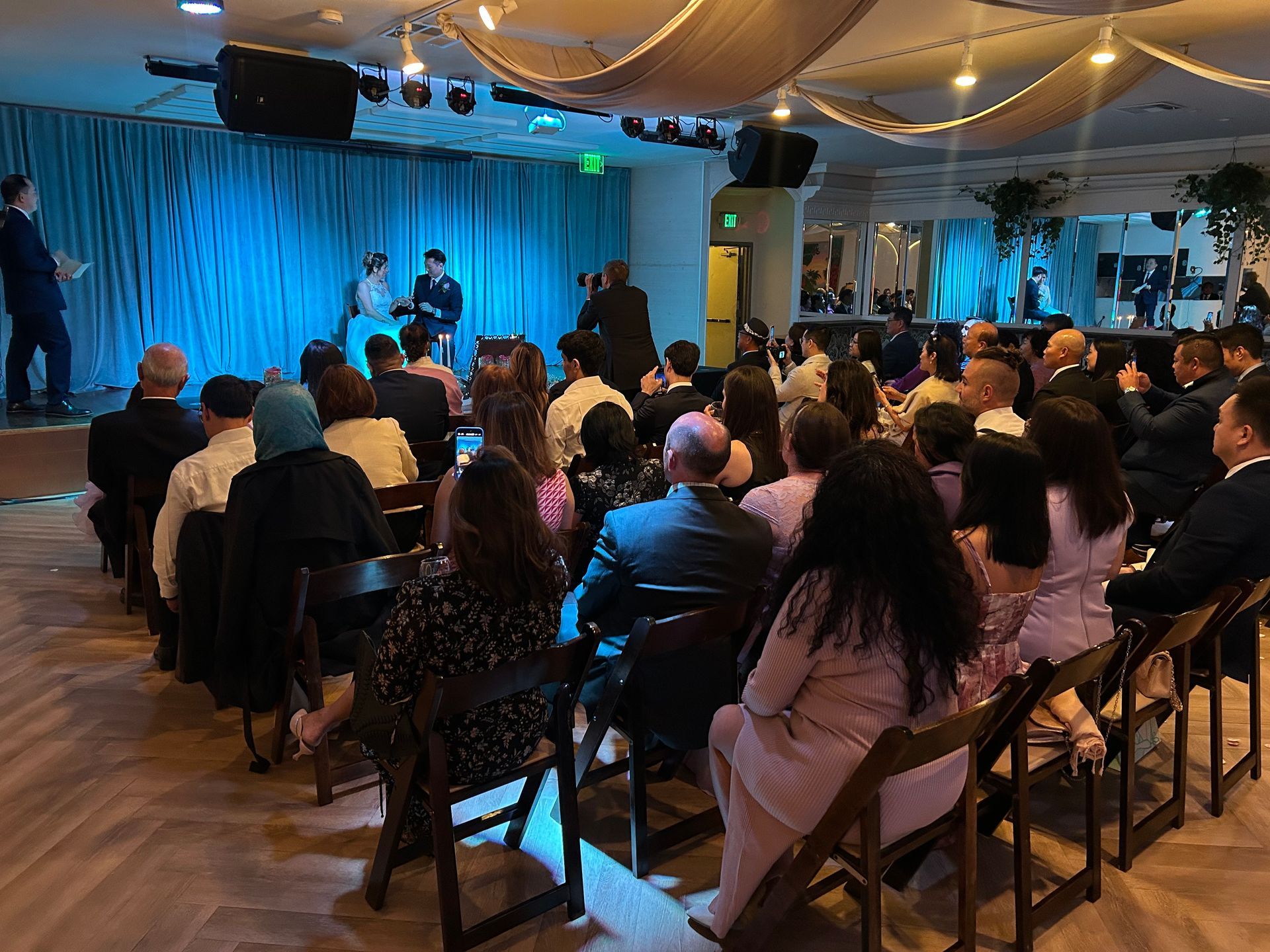 Audience seated at a live performance on a small stage with blue-lit drapes and a speaker backdrop