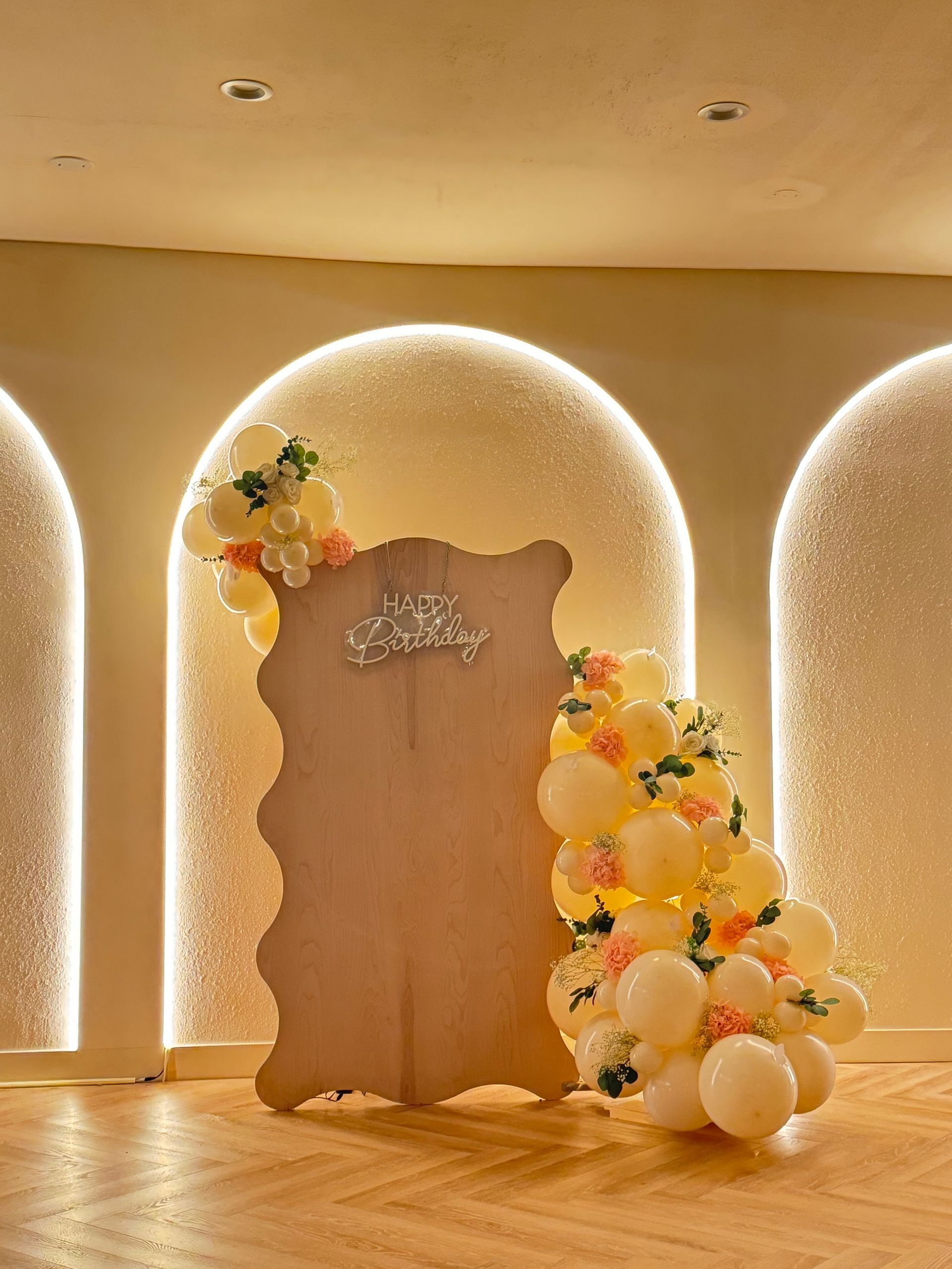 Elegant event backdrop with a wooden board, floral accents, and pastel balloon arrangement in a softly lit archway.