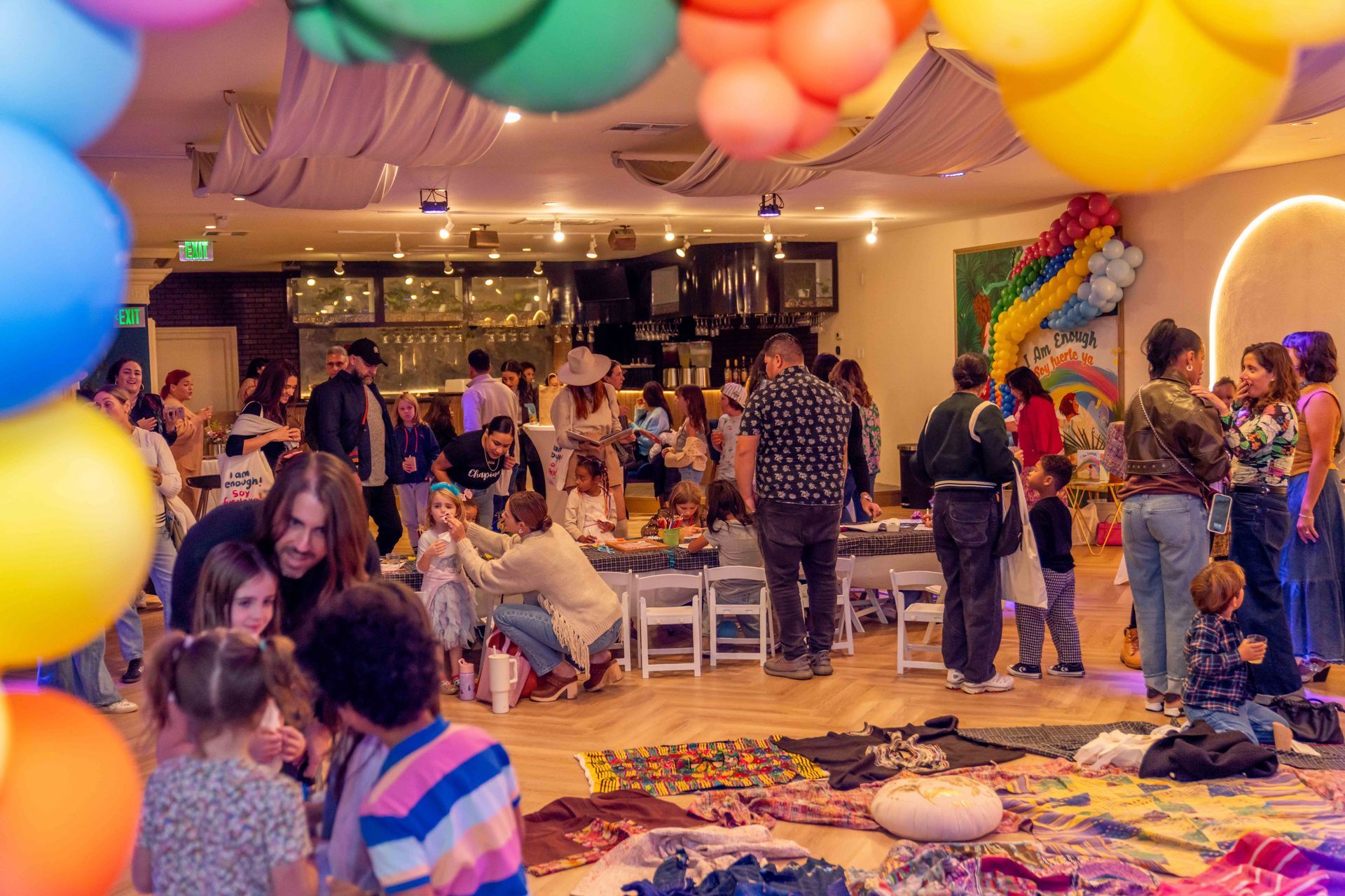 Colorful indoor party with balloons, people mingling, and children playing on a decorated floor
