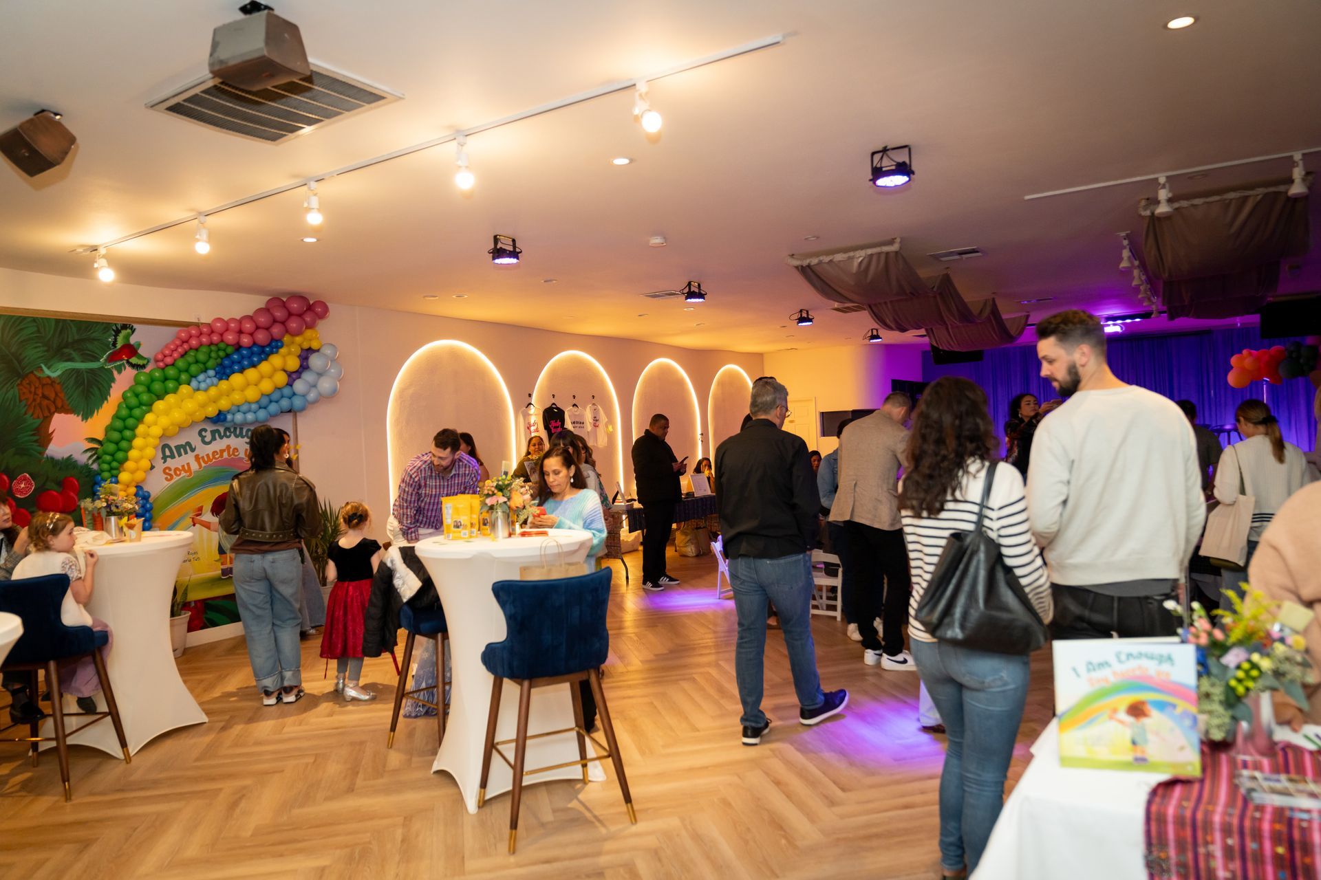 Guests mingle in a warmly lit banquet hall with colorful wall decor and white tablecloths.