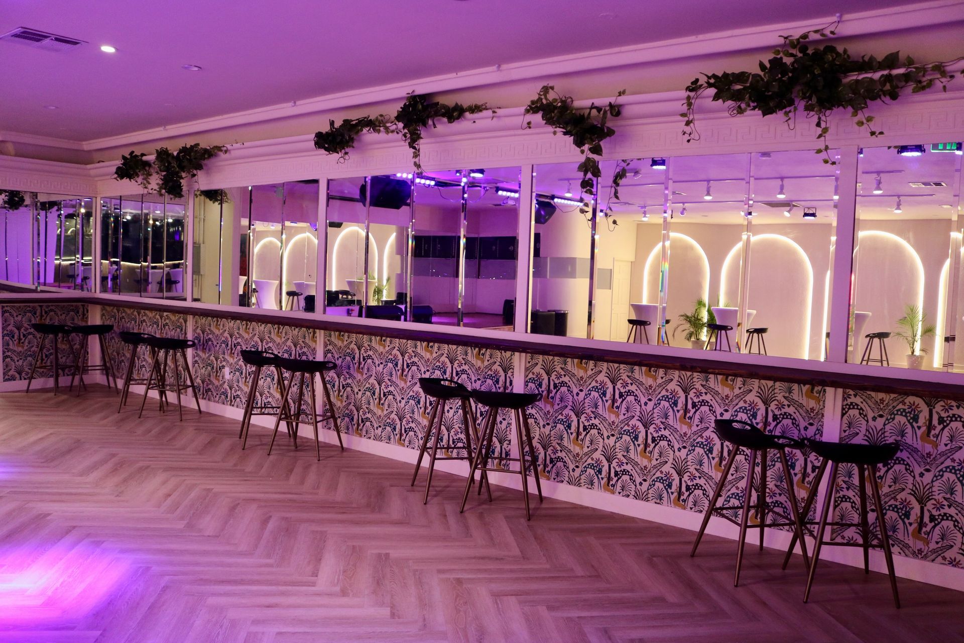 Purple-lit modern bar interior with mirrored wall, high stools, and hanging greenery