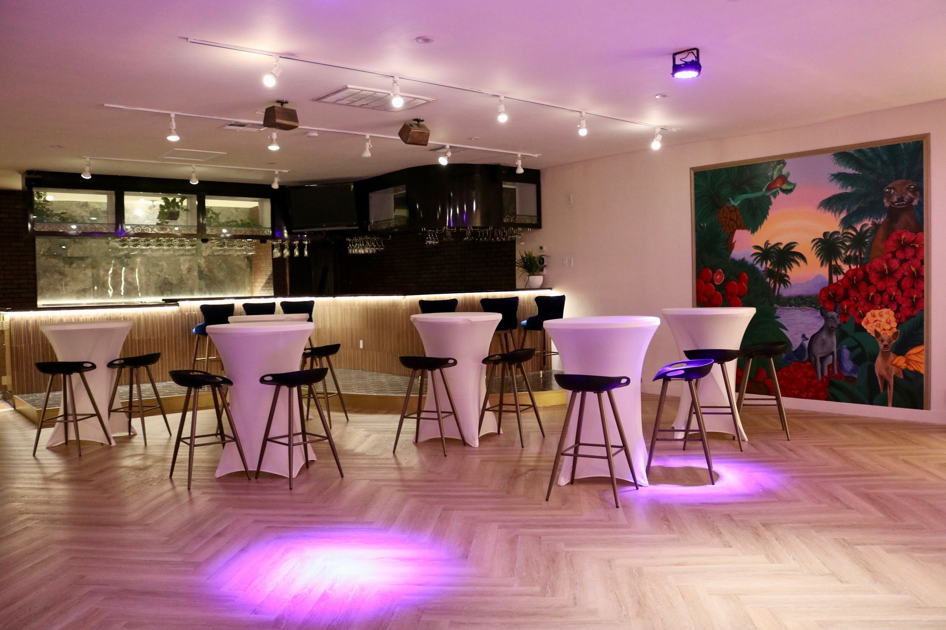 Modern lounge with high tables, stools, pink lighting, and a colorful mural on the wall.