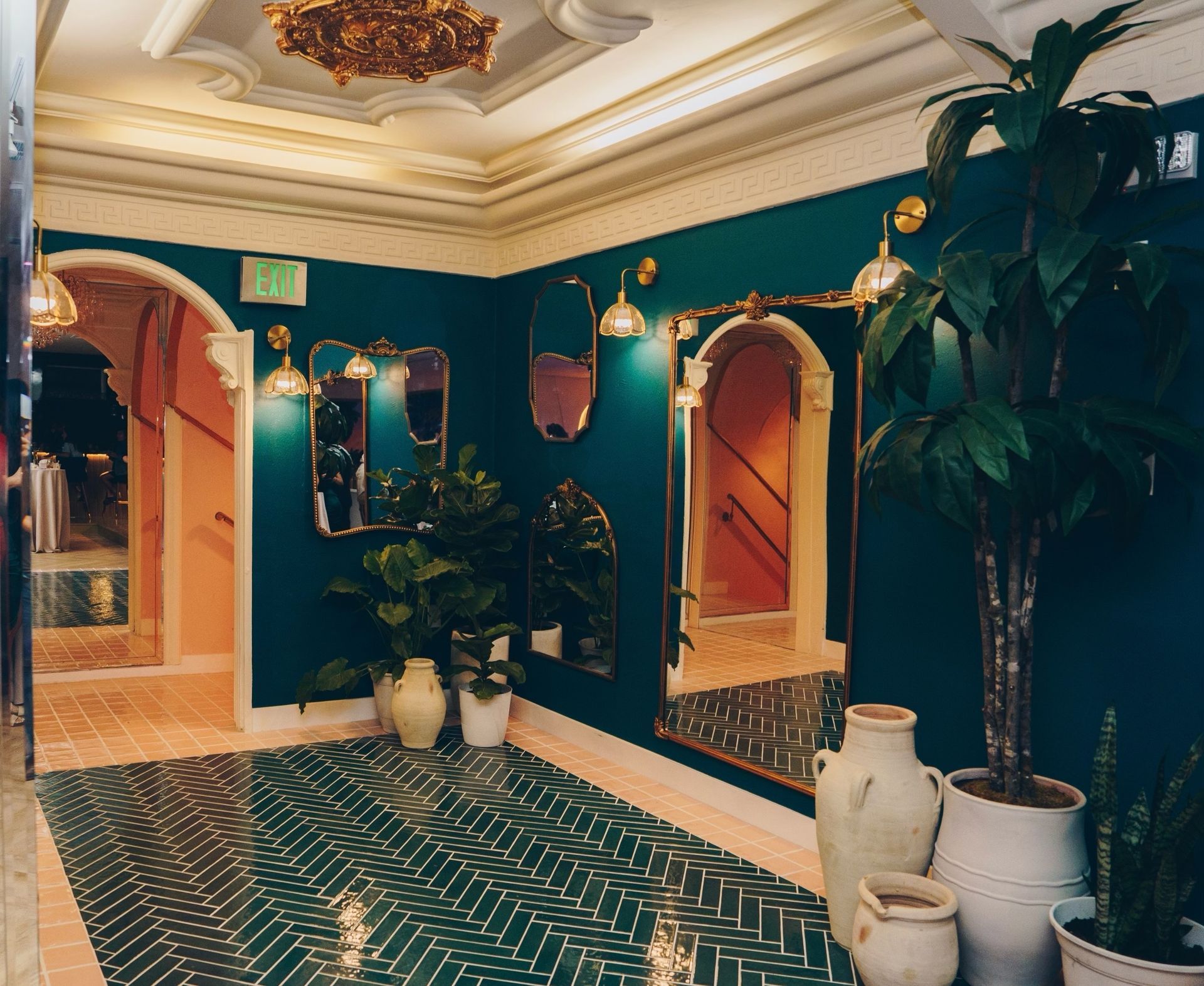 Elegant teal hallway with gold mirrors, ornate ceiling, patterned floor, and large potted plants
