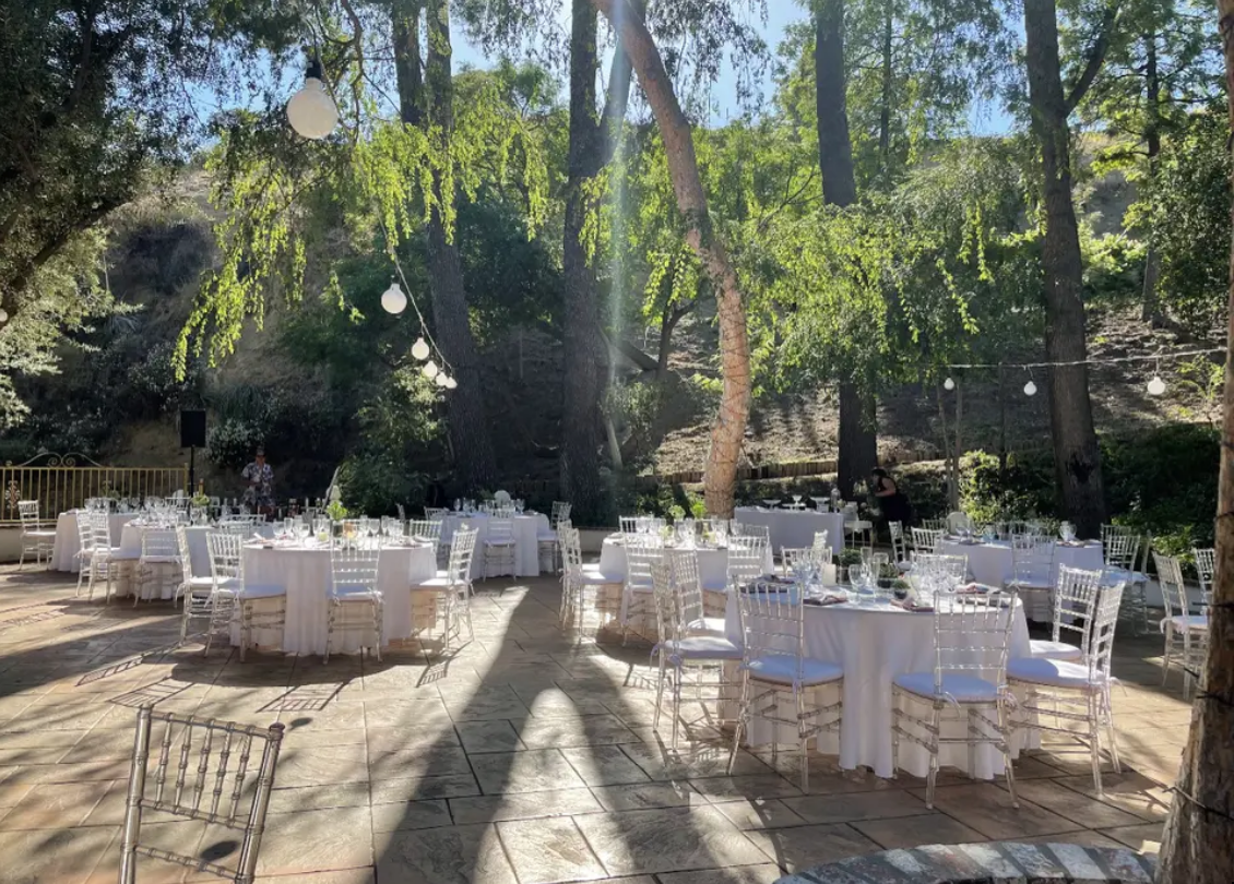 Outdoor wedding reception setup with white tables and chairs under trees in a sunlit garden
