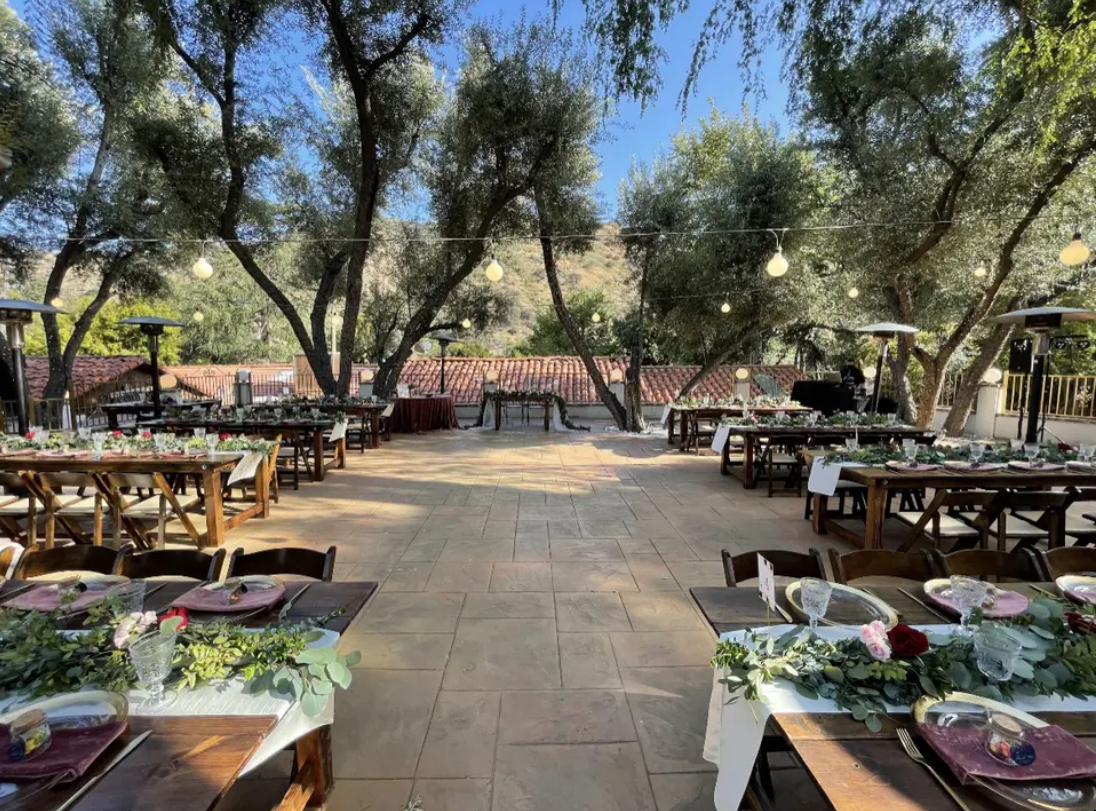 Outdoor dining area under trees with long wooden tables set for a meal on a stone patio