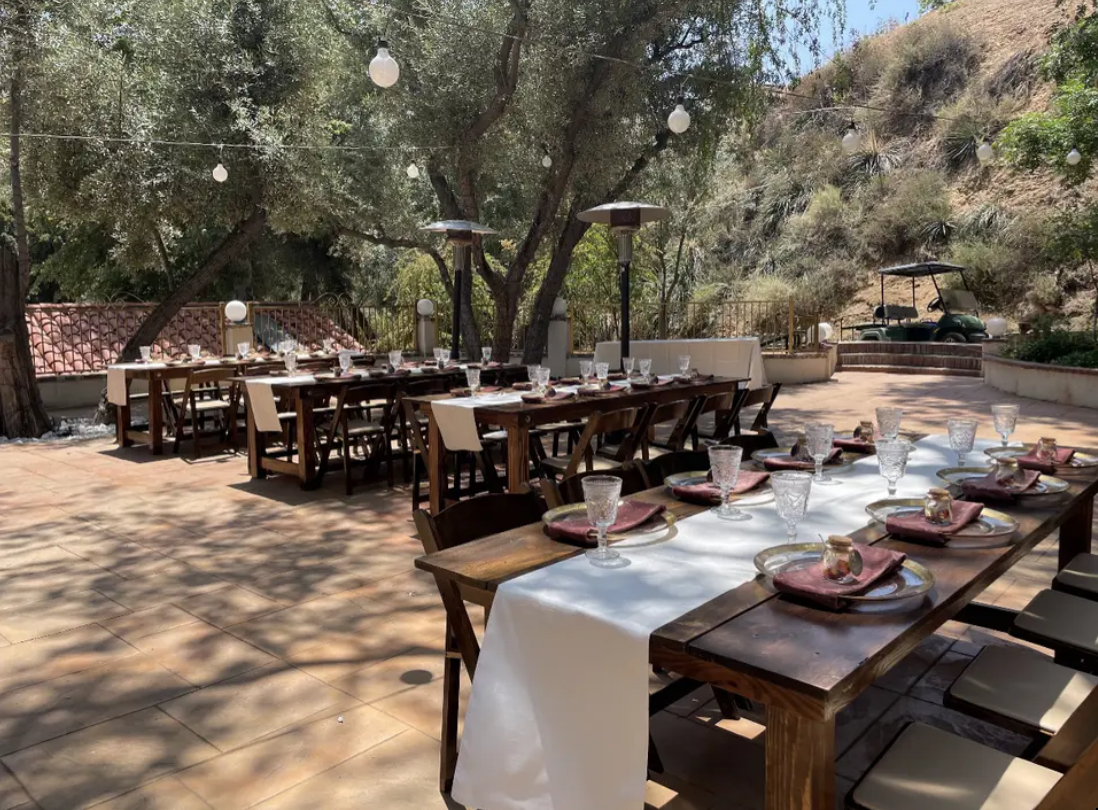 Outdoor dining area with long wooden tables set with white cloths under trees in a sunlit canyon.