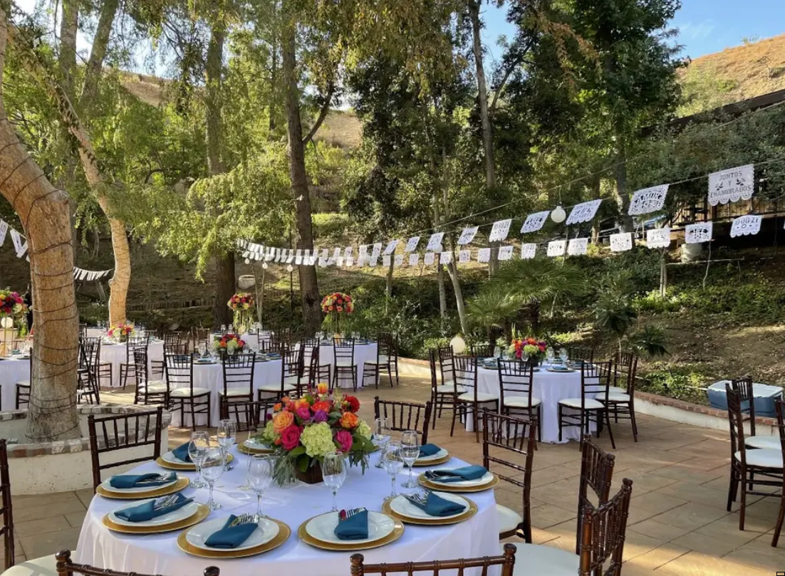 Outdoor banquet table set with blue plates, floral centerpiece, and stringed photos under trees