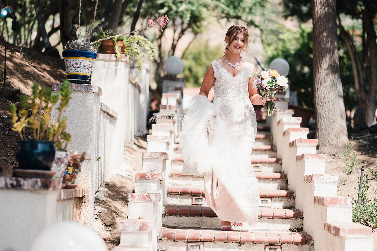 Bride in a white dress walking down outdoor steps with a bouquet in a garden setting