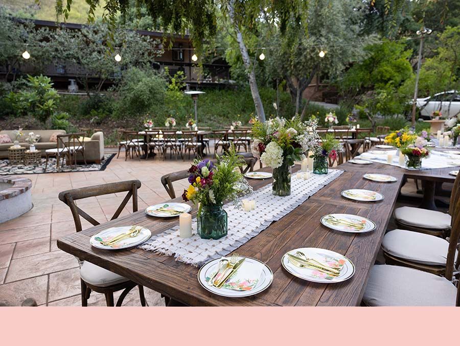 Outdoor dining tables set with white plates and flower centerpieces on a patio surrounded by greenery.
