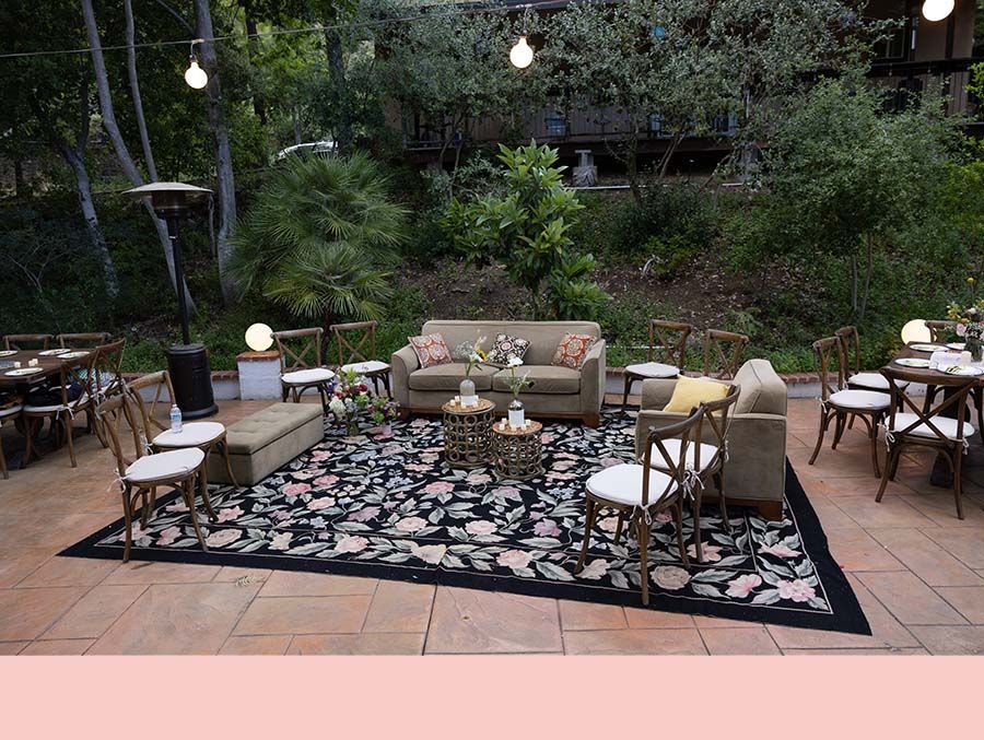 Outdoor patio seating area with couches, chairs, and string lights on a patterned rug, surrounded by greenery