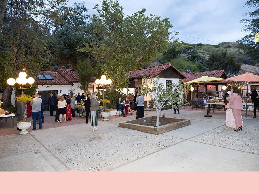 Outdoor evening gathering with people under lit trees, small buildings, and a paved courtyard