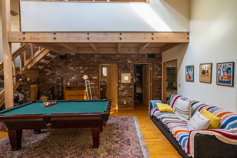 Rec room with pool table, colorful couch, patterned rug, and loft overlooking brick wall and wooden staircase