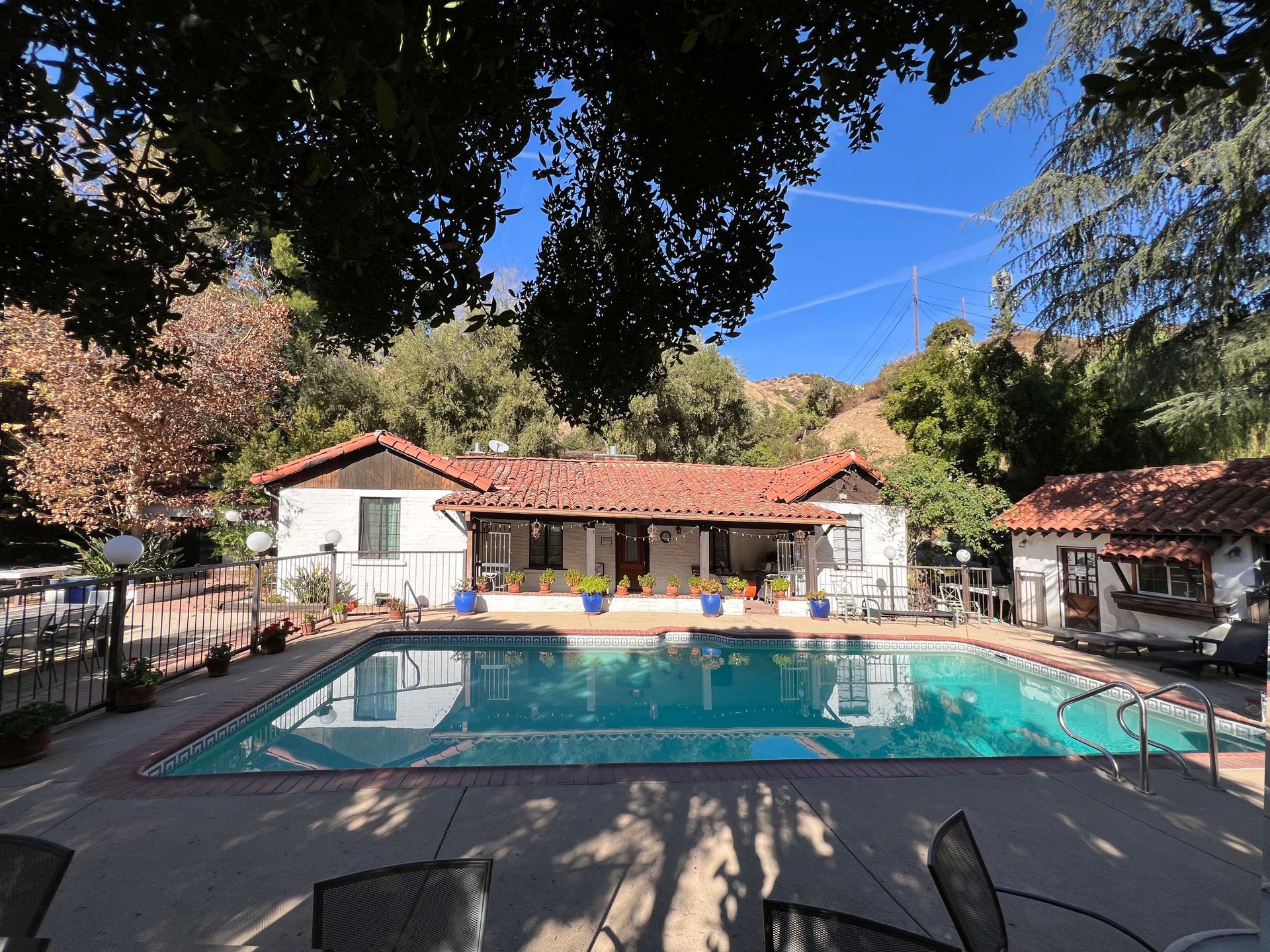 Backyard swimming pool with lounge chairs and a white house under trees, viewed from a shaded patio
