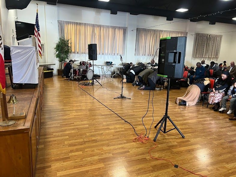 A group of people are sitting in a large room with a wooden floor.
