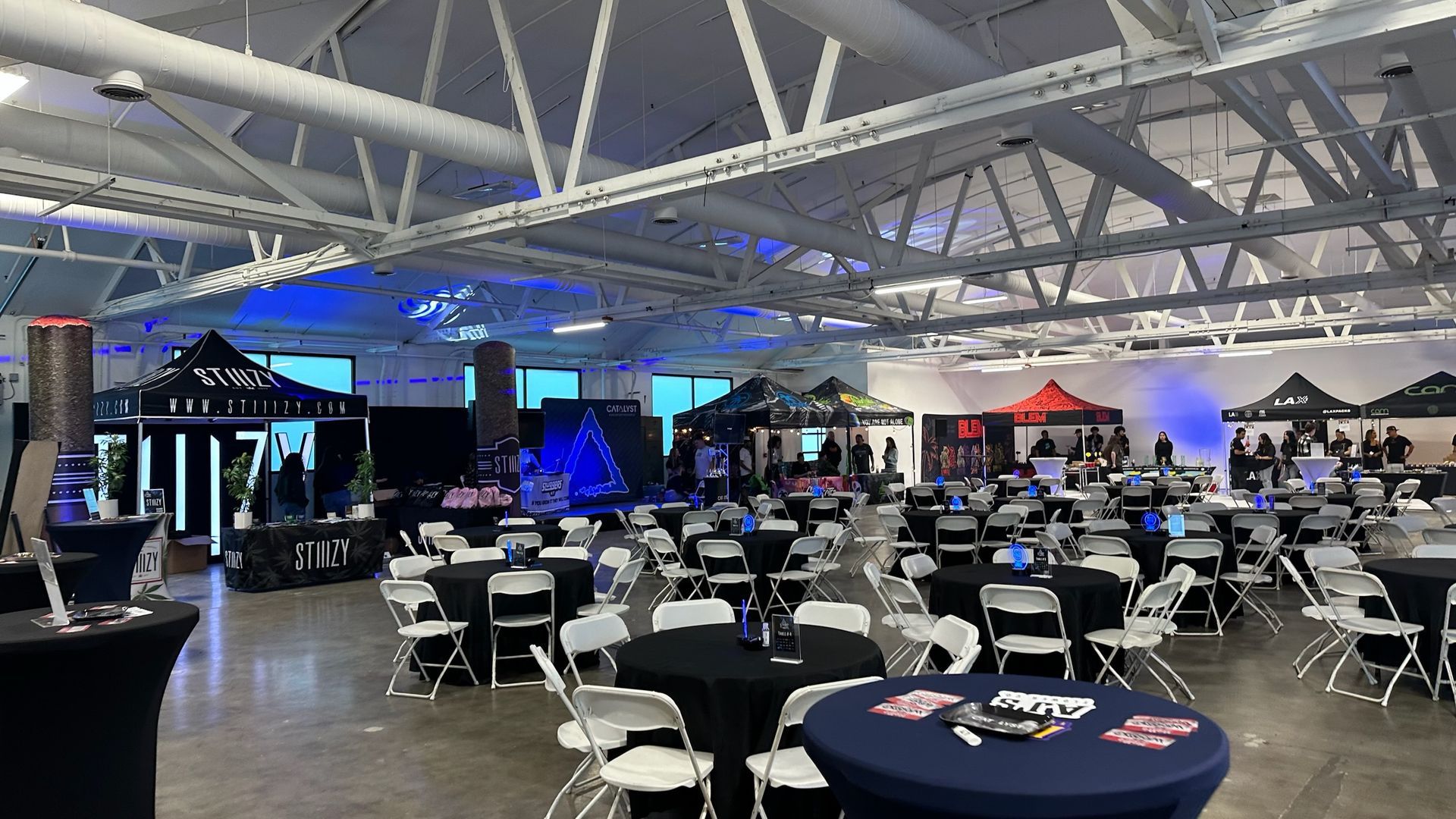 An indoor event space set up with rows of round tables with black tablecloths and white folding chairs under a high roof.