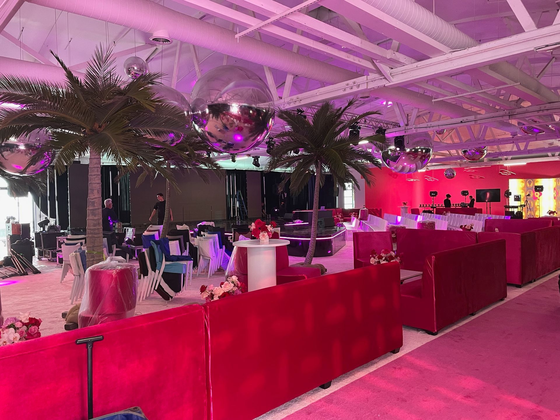A bright pink event space with palm trees, tiered red lounge seating, and disco balls hanging from the industrial ceiling.