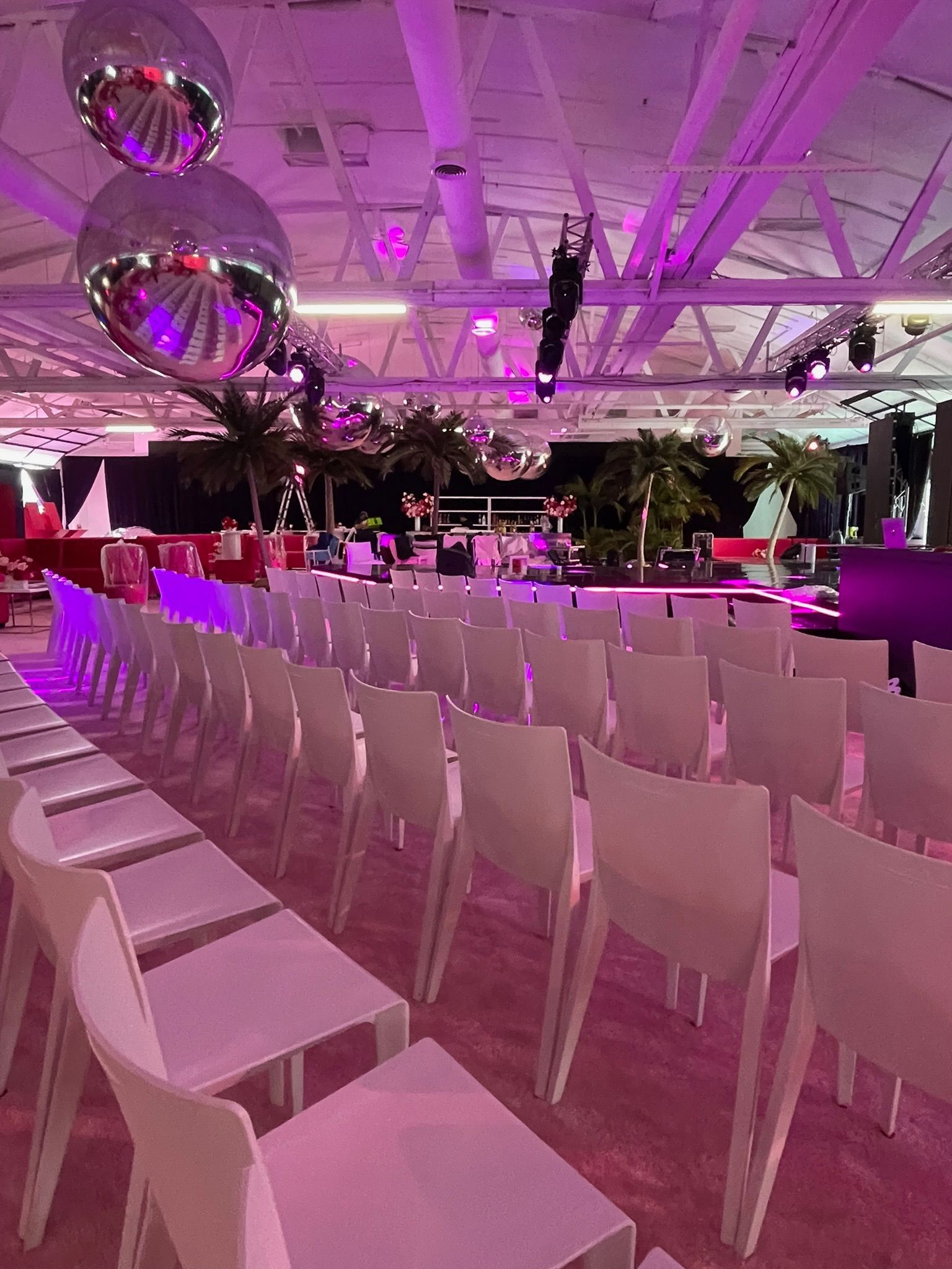 Rows of white chairs face a stage area in a spacious venue with disco balls, pink lighting, and palm trees.