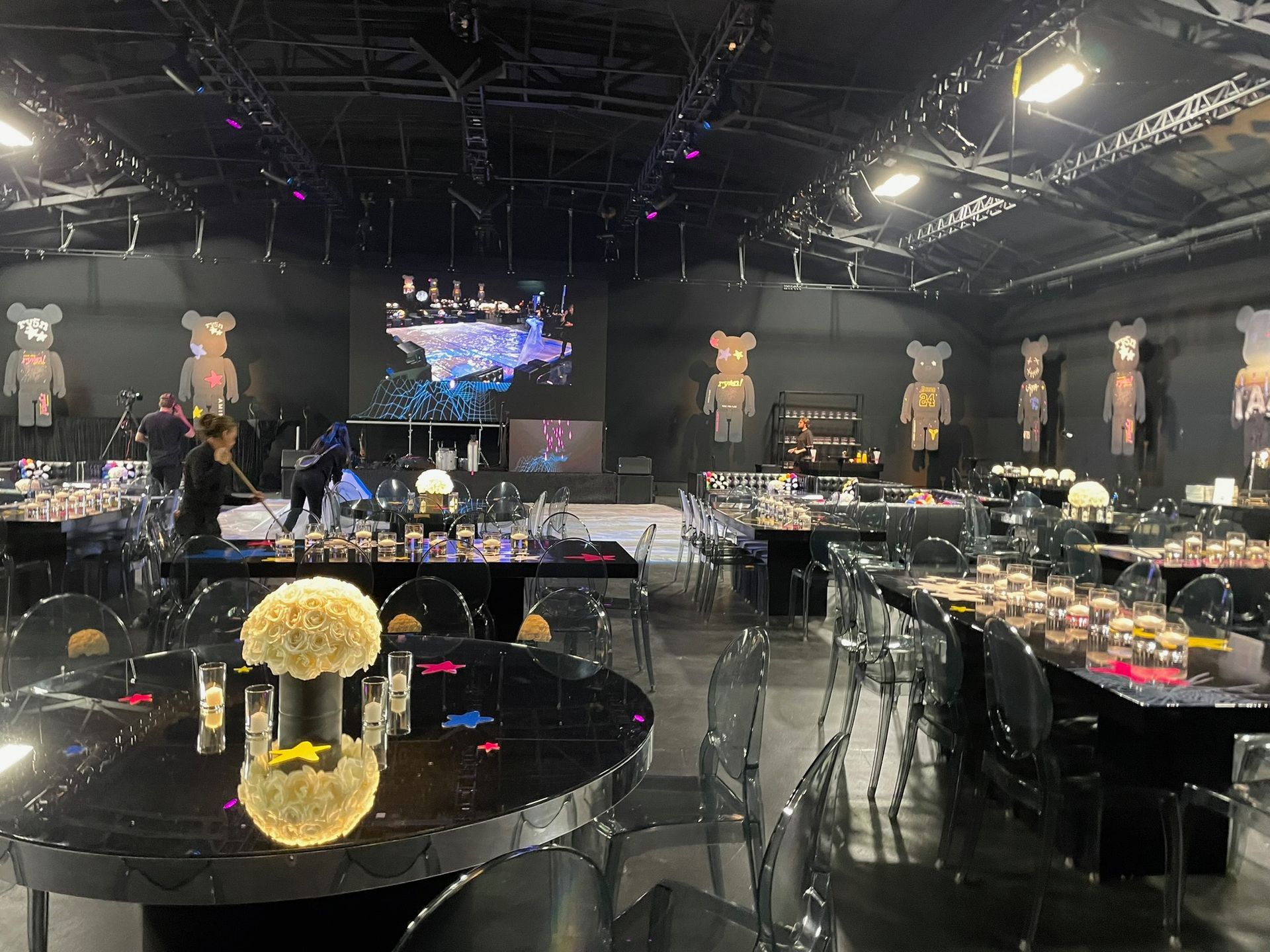 A dimly lit event space featuring round tables with floral centerpieces, decorated with several large Bearbrick figures.