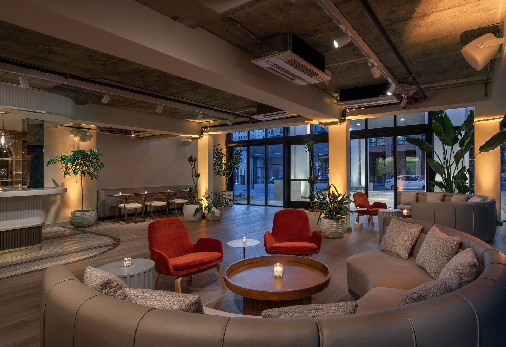 A modern hotel lobby featuring a curved gray sofa, two red armchairs, a wooden coffee table, and large potted plants.