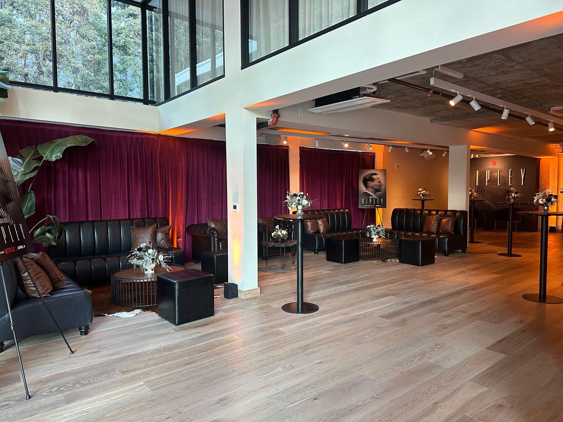 A modern, airy lounge with purple curtains, leather sofas, small cocktail tables, and light wooden floors.