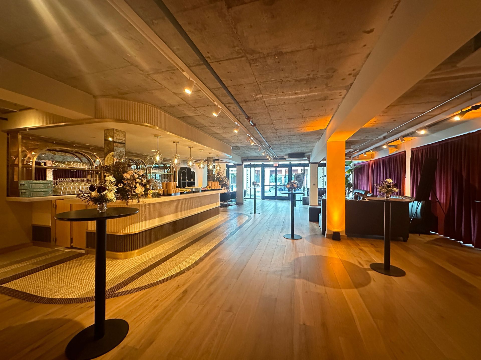 Modern event space with a bar, wood floors, exposed concrete ceiling, and tall cocktail tables under warm lighting.