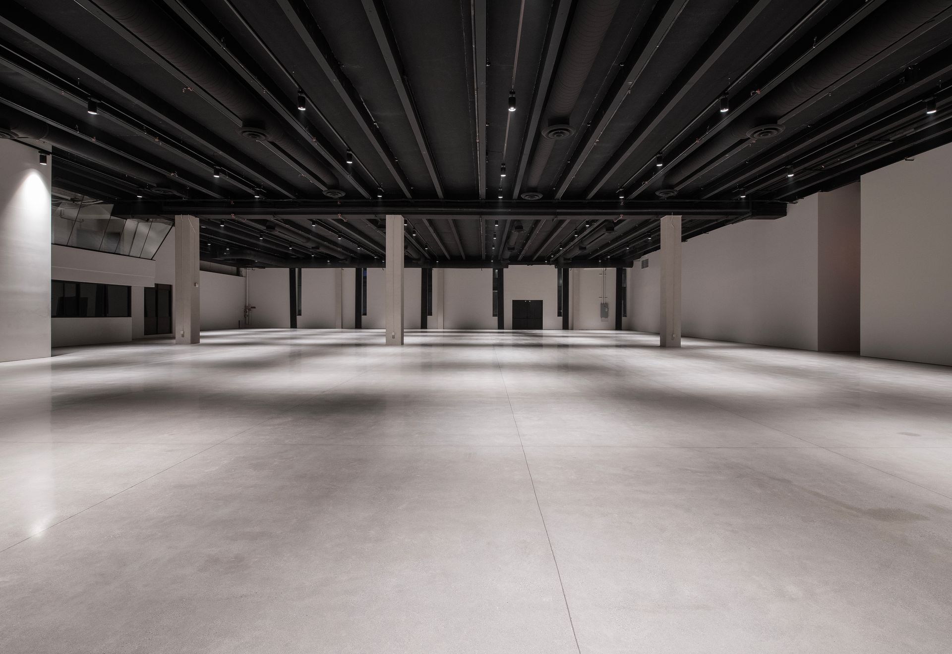 A wide, empty industrial-style event space with polished concrete floors, white walls, and a black open-beam ceiling.