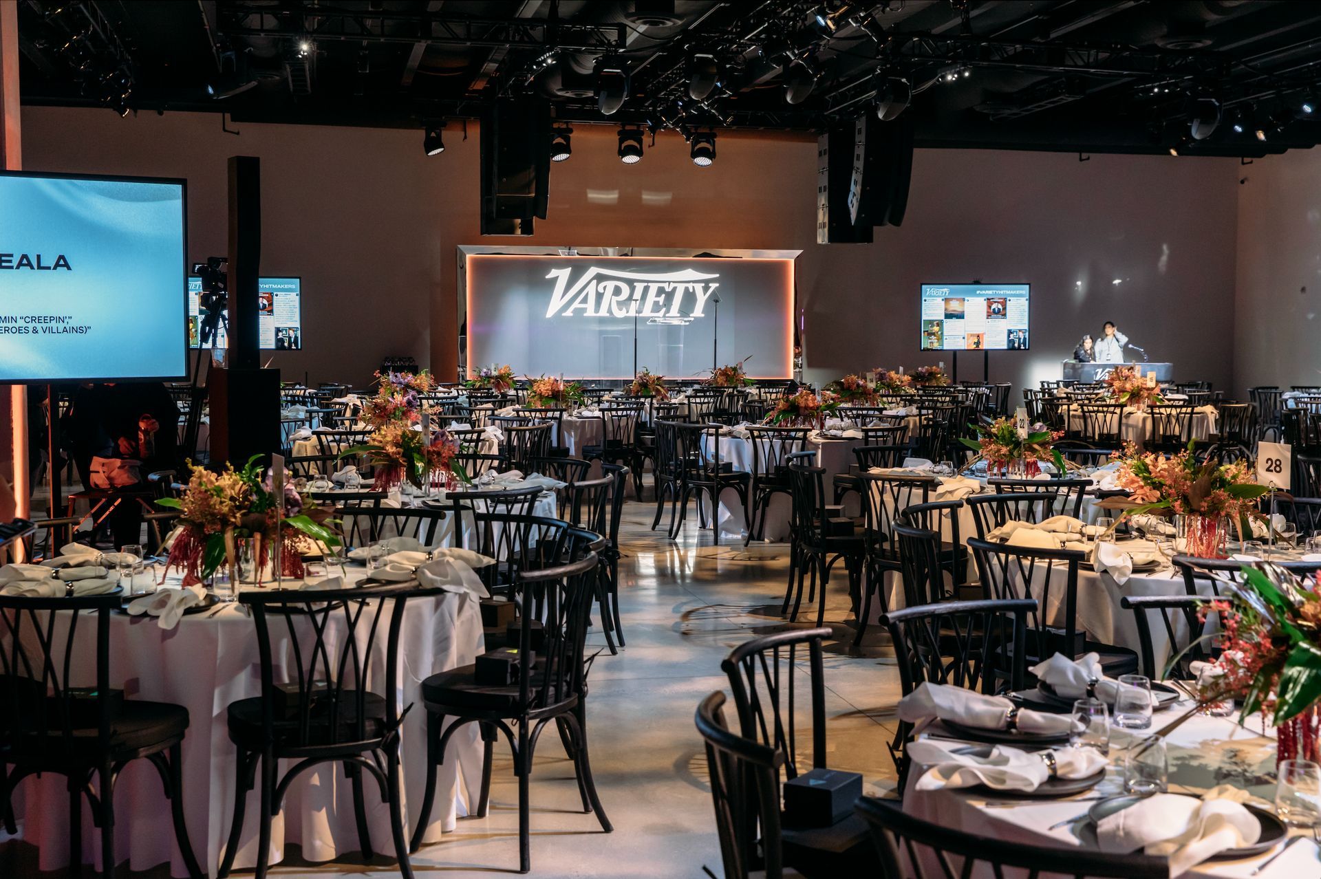 An event hall set for a banquet, featuring round tables with white tablecloths, floral centerpieces, and Variety branding.