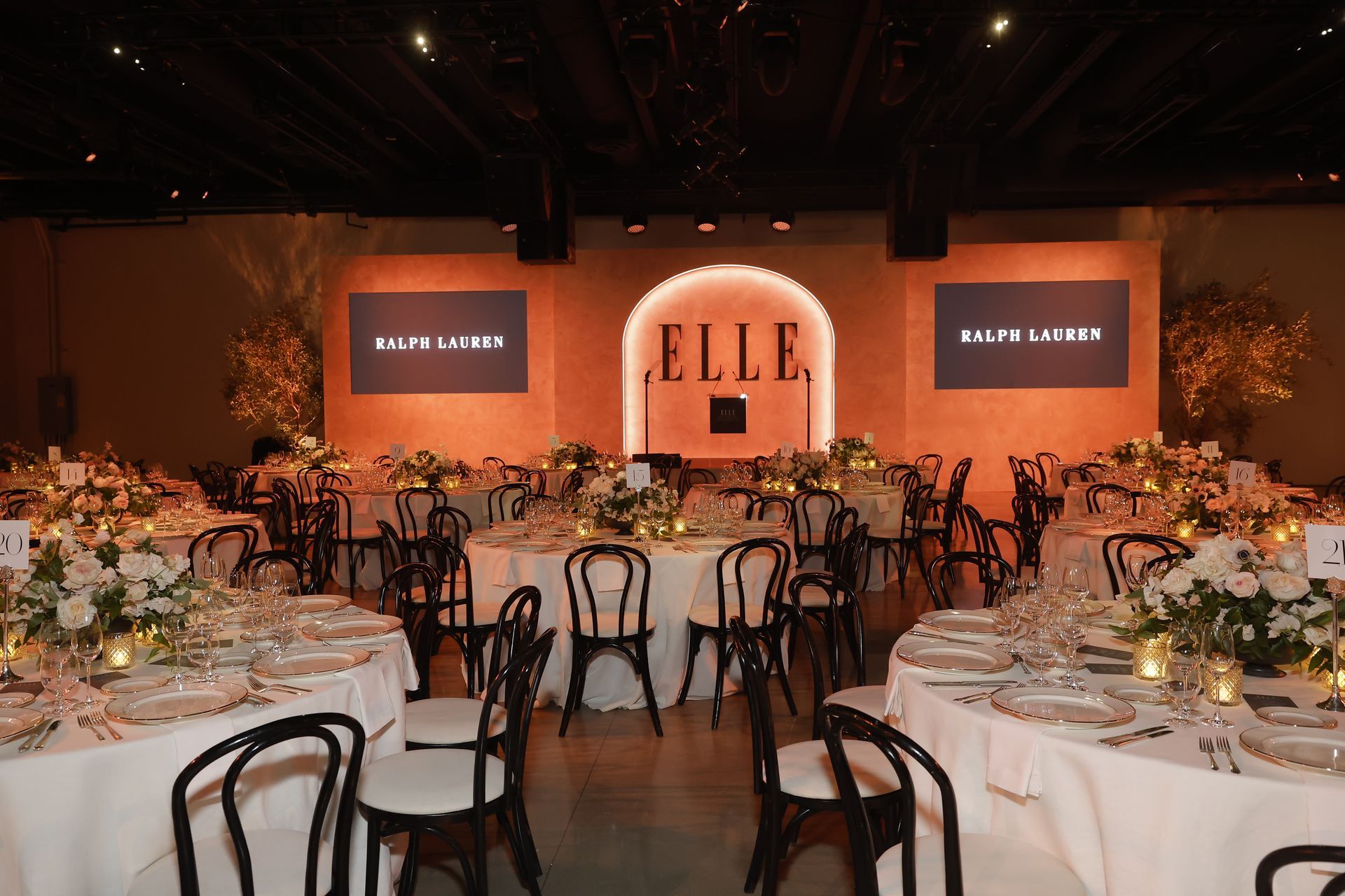 A banquet hall set for a Ralph Lauren and ELLE event with white-clothed tables, chairs, and a branded stage backdrop.