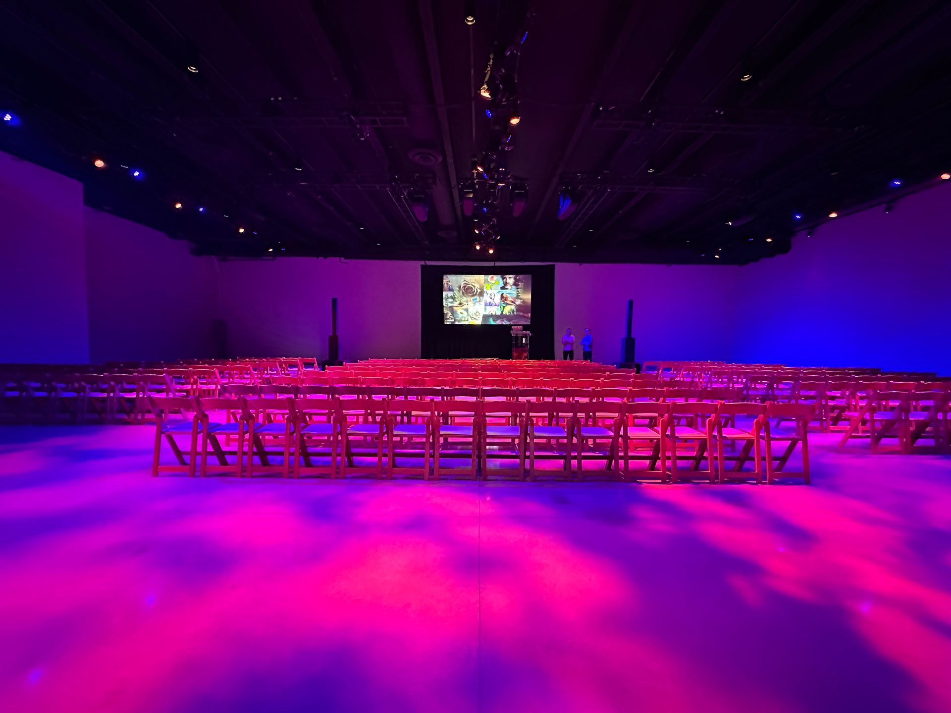 An empty event space with rows of chairs facing a screen, illuminated by vibrant pink and blue floor lighting.
