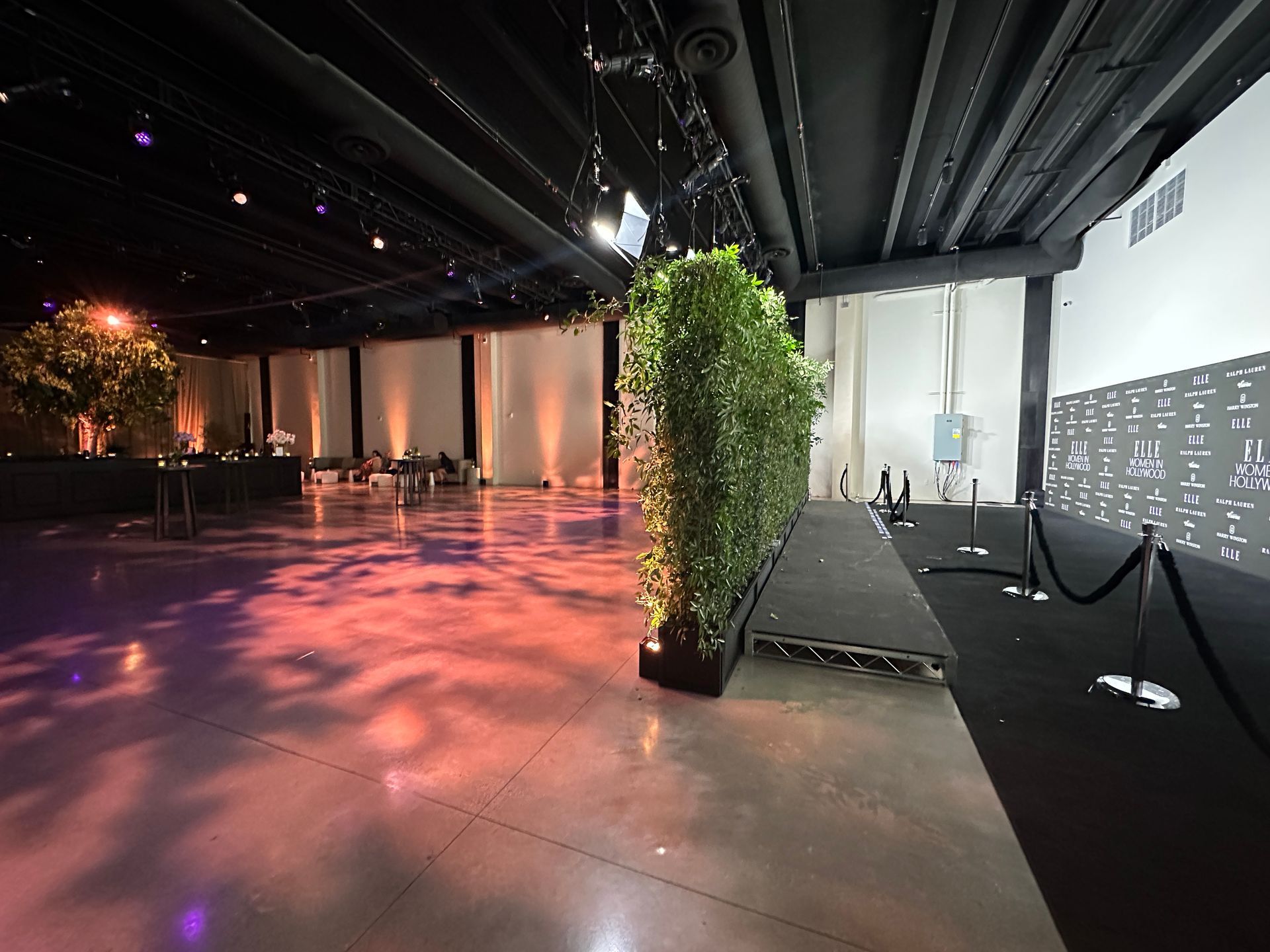 A spacious event hall with polished floors, a hedge partition on a low stage, and a step-and-repeat backdrop.