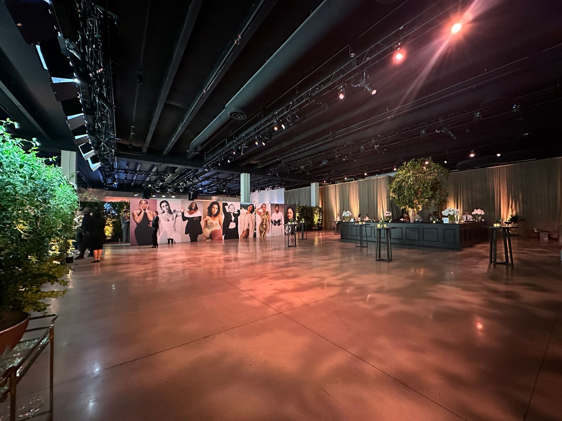 A spacious, modern event hall with polished floors, warm lighting, a photo wall, and green decor under a black ceiling.