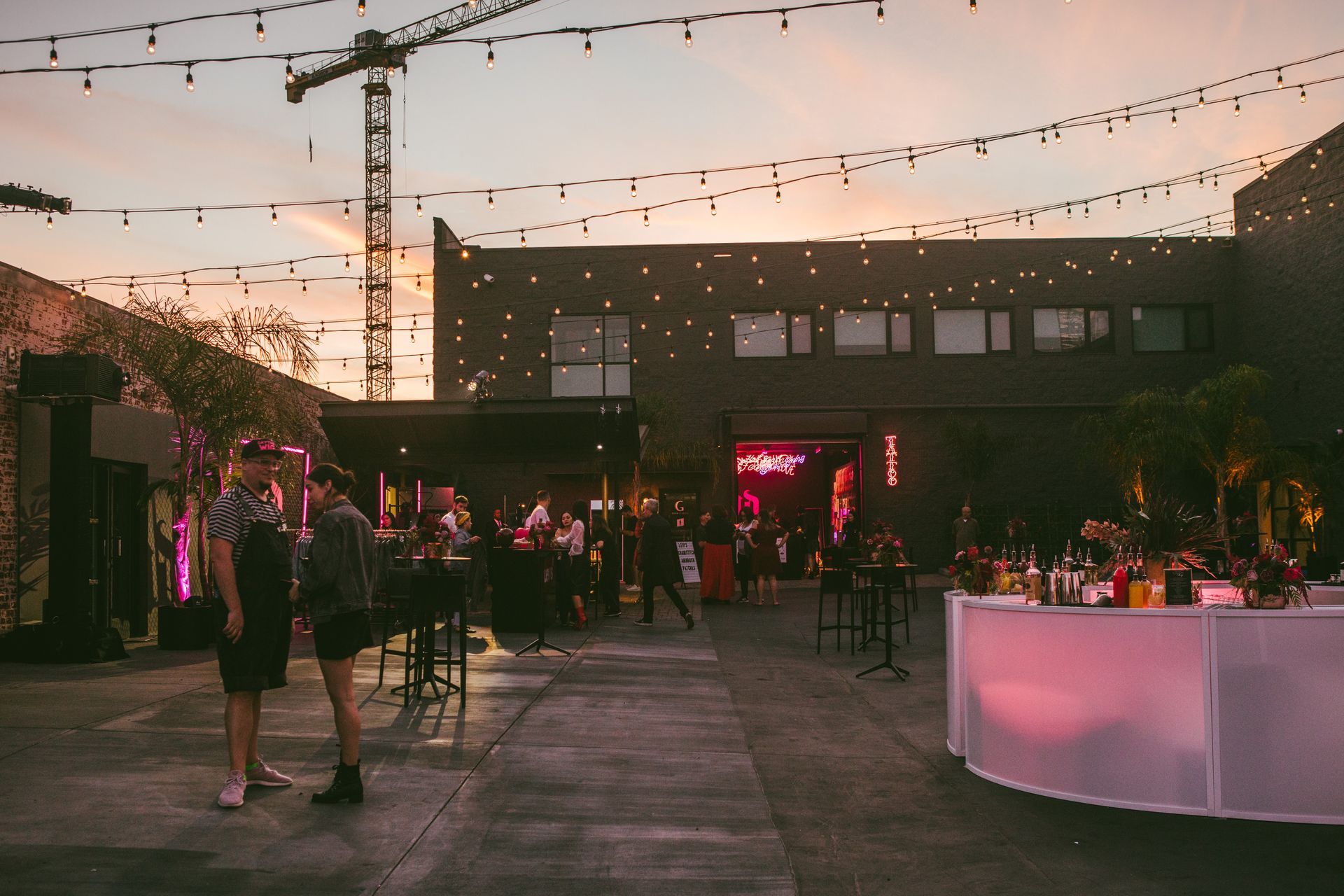 People socialize at an outdoor evening event with string lights, a neon sign, and a glowing bar under a construction crane.