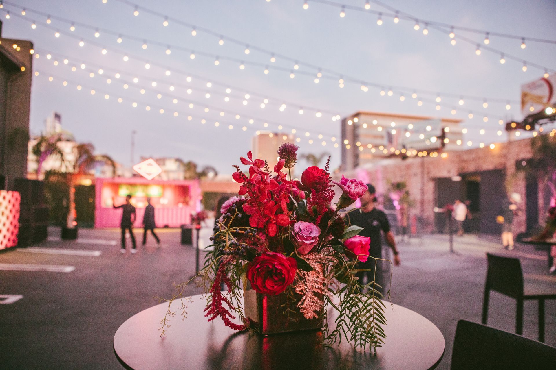 A vibrant floral arrangement sits on a table at an outdoor event venue decorated with string lights at twilight.
