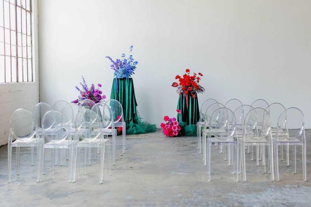 Empty rows of transparent chairs face two floral arrangements on green pedestals in a minimalist, industrial-style room.