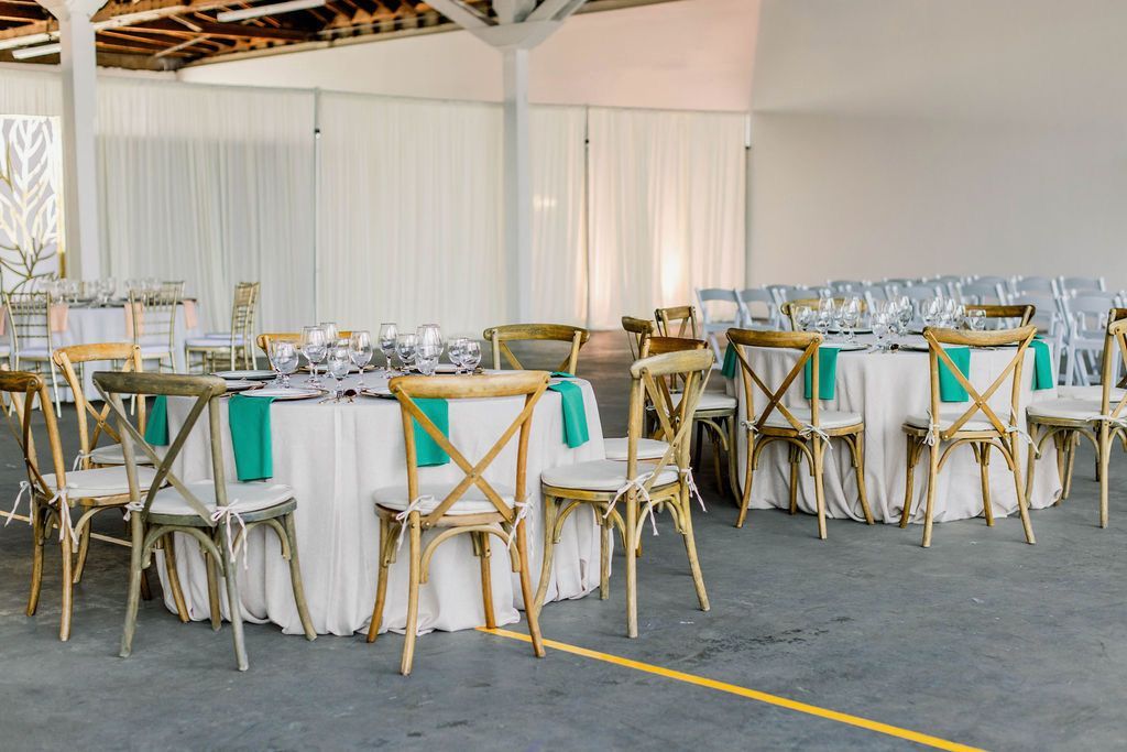 Round event tables with white linens, teal napkins, and light-wood cross-back chairs arranged in a large indoor venue.