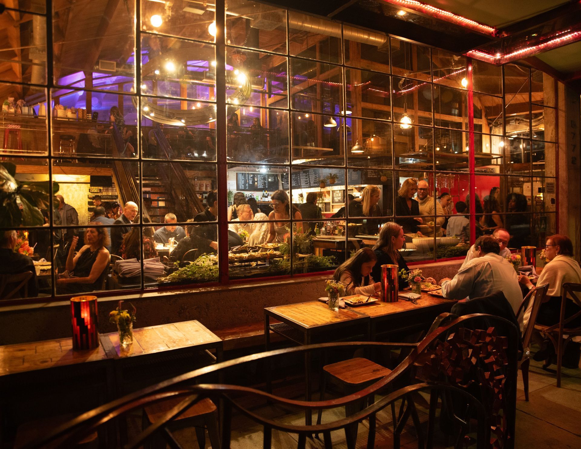 Patrons dine in an outdoor patio area with a view of a dimly lit, warm-toned restaurant interior through large windows.
