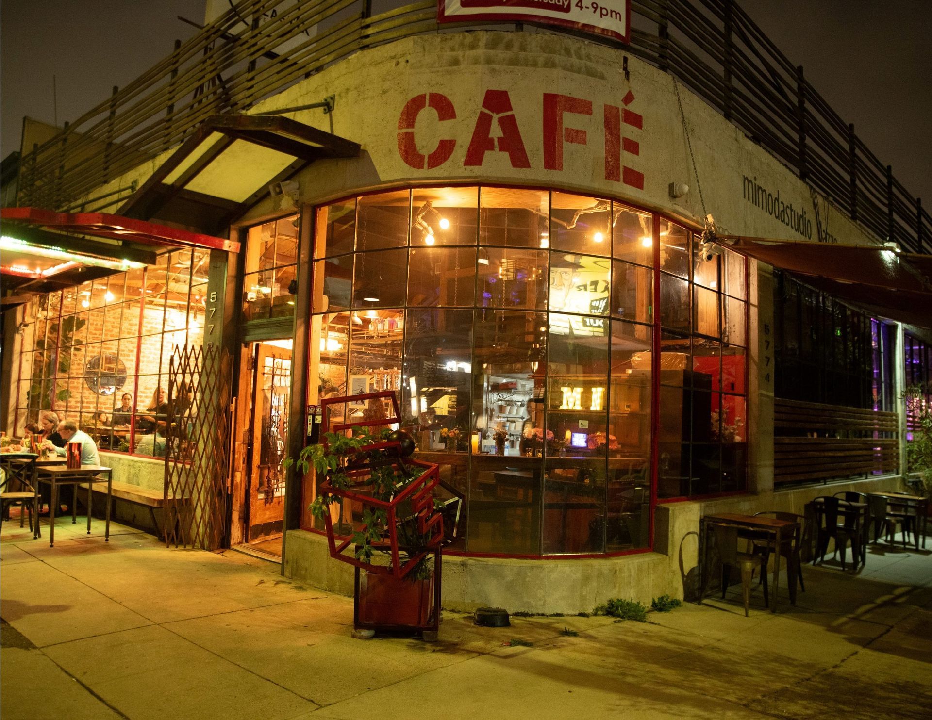 A corner cafe at night with large, multi-paned windows, warm interior lighting, and outdoor sidewalk seating.