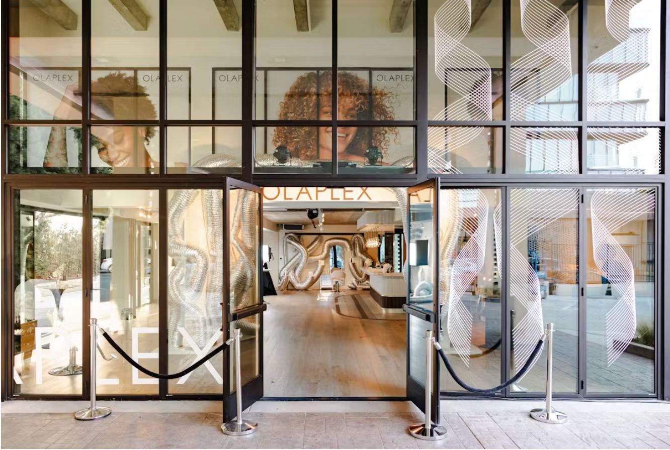 An Olaplex storefront with floor-to-ceiling glass windows and doors, featuring interior branding and rope stanchions.