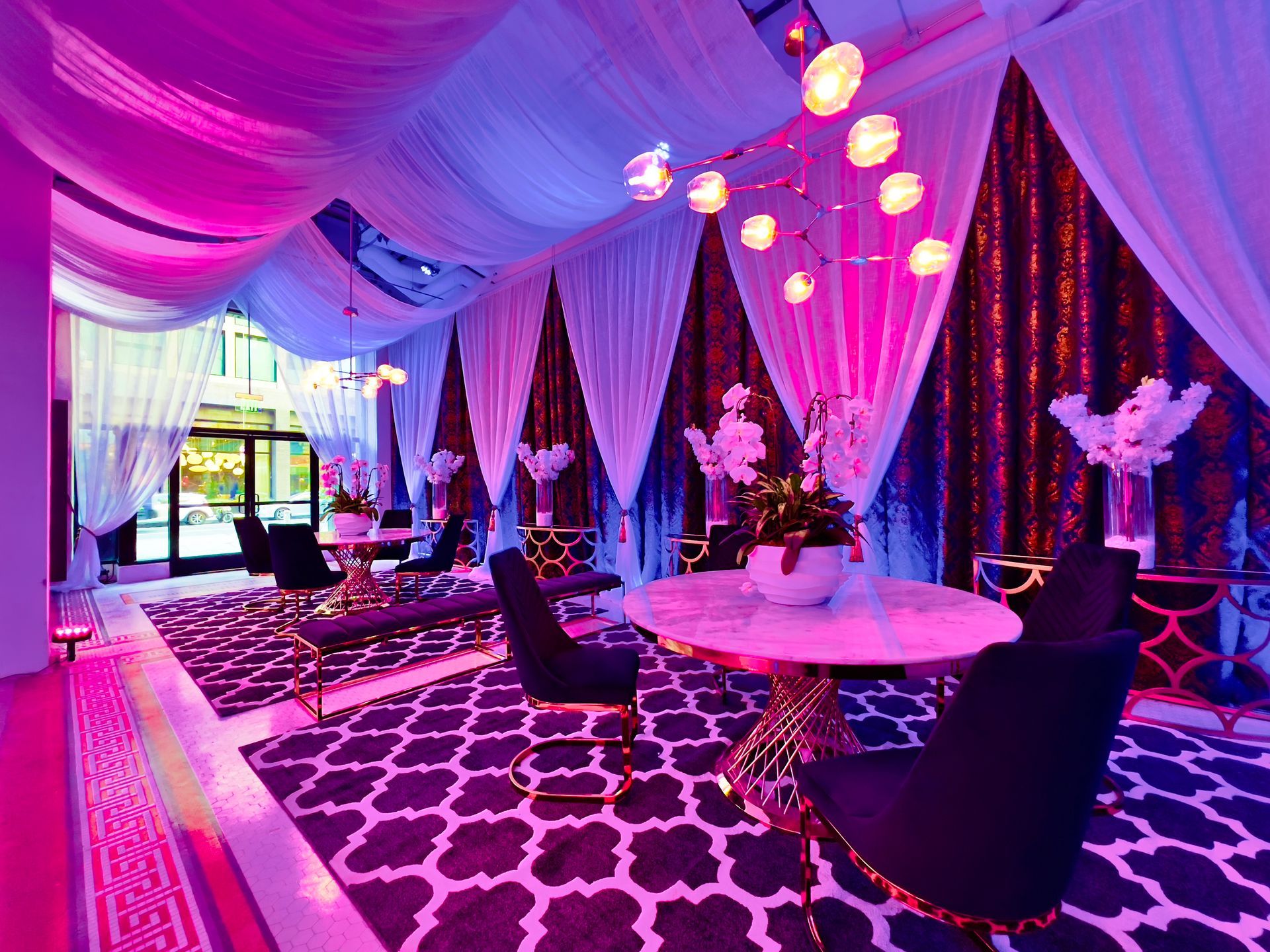 Lavender-lit event lounge with draped ceiling, round tables, and modern chairs on patterned carpet