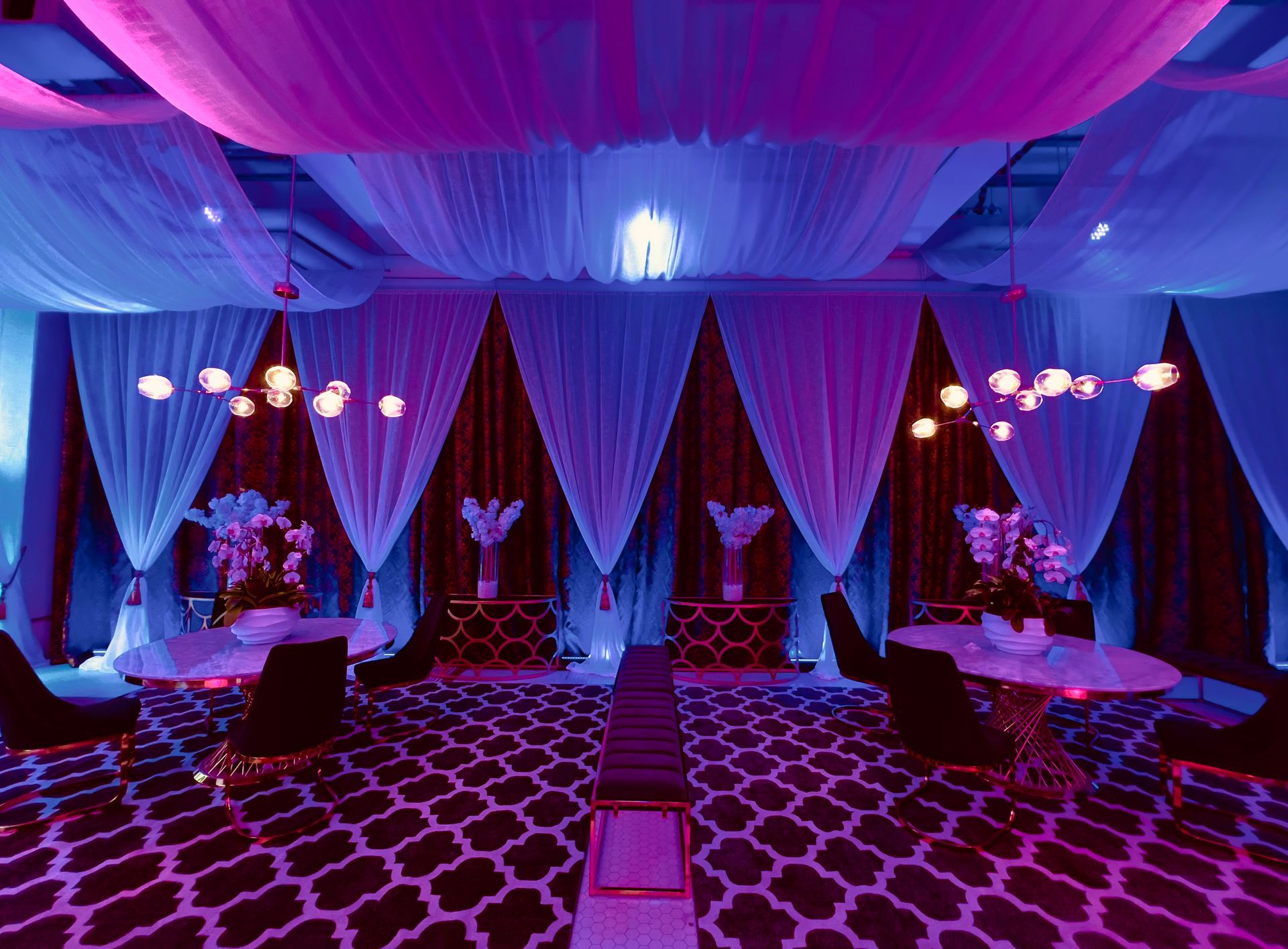 Elegant event hall with purple-blue lighting, draped curtains, hanging lights, and round tables on a patterned floor