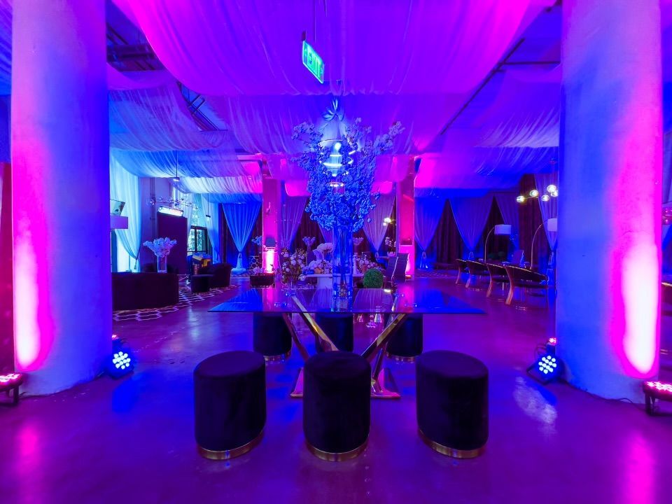 Elegant event venue with purple-blue uplighting, draped columns, and a central floral arrangement with stools