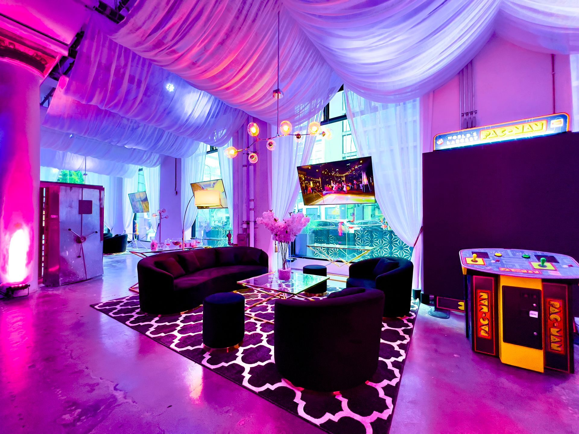 Neon-lit arcade lounge with black couches, glowing floor decor, and game cabinets under pink draped ceiling