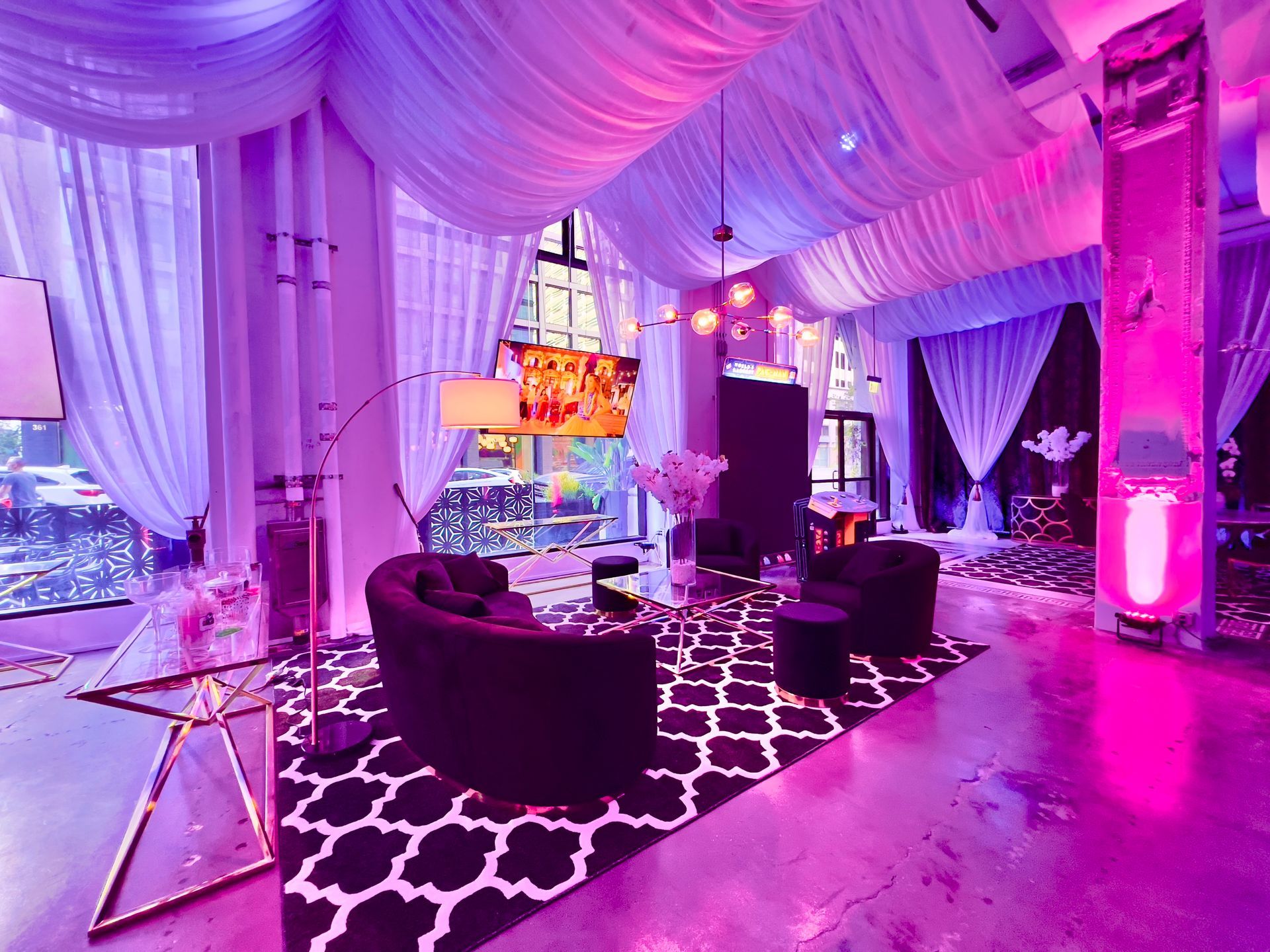 Elegant lounge with purple drapes, pink lighting, black sofas, and patterned rugs in a decorated event space