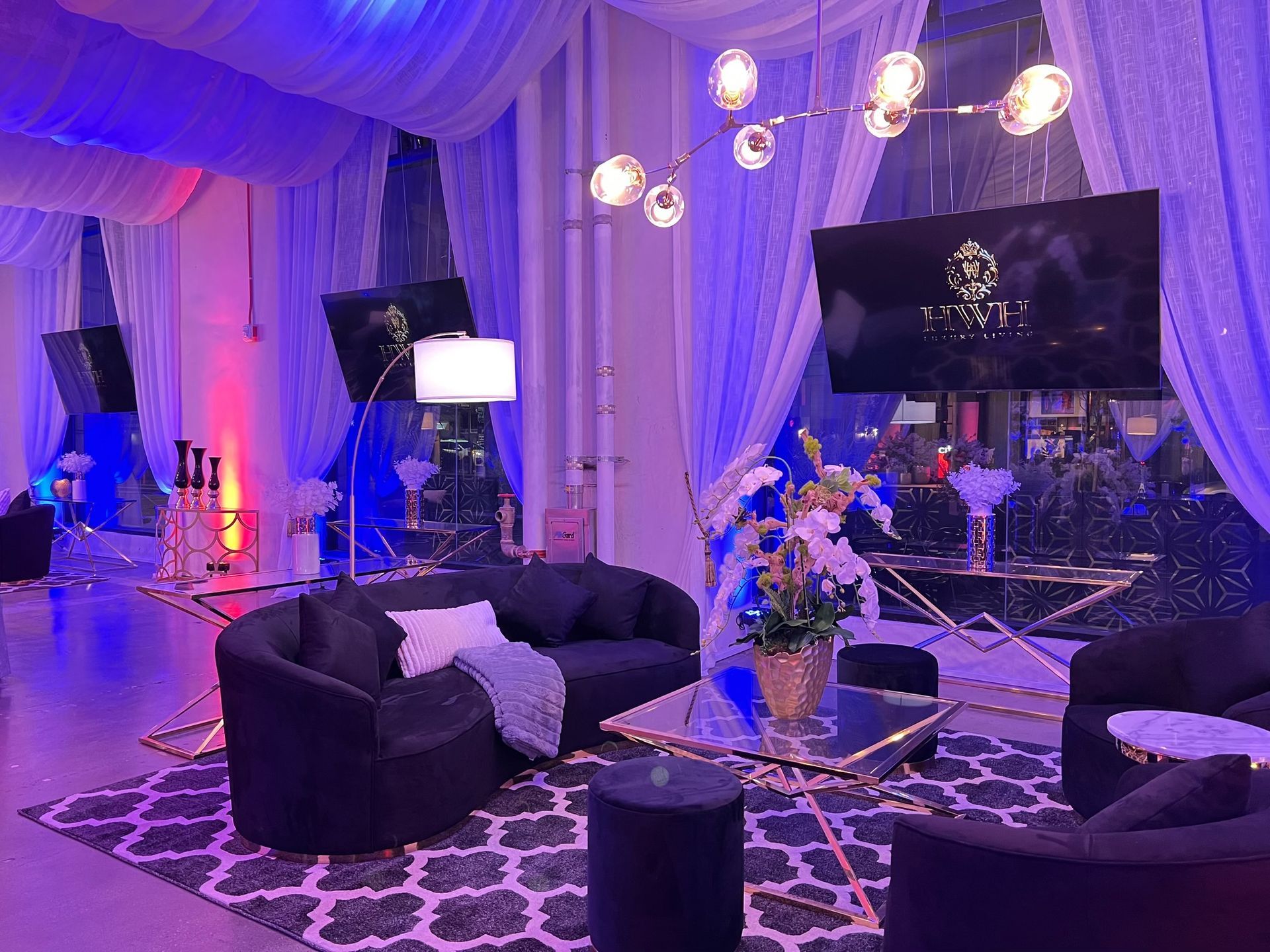 Elegant lounge with purple lighting, black sofas, patterned rug, floral decor, and wall-mounted TVs