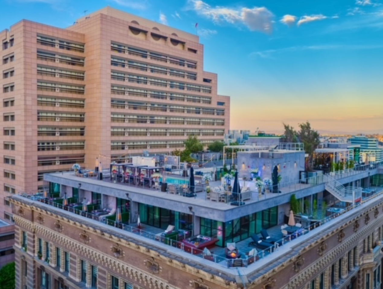 A rooftop terrace with lounge seating and a pool, set against a tall, modern tan building under a blue sky at sunset.