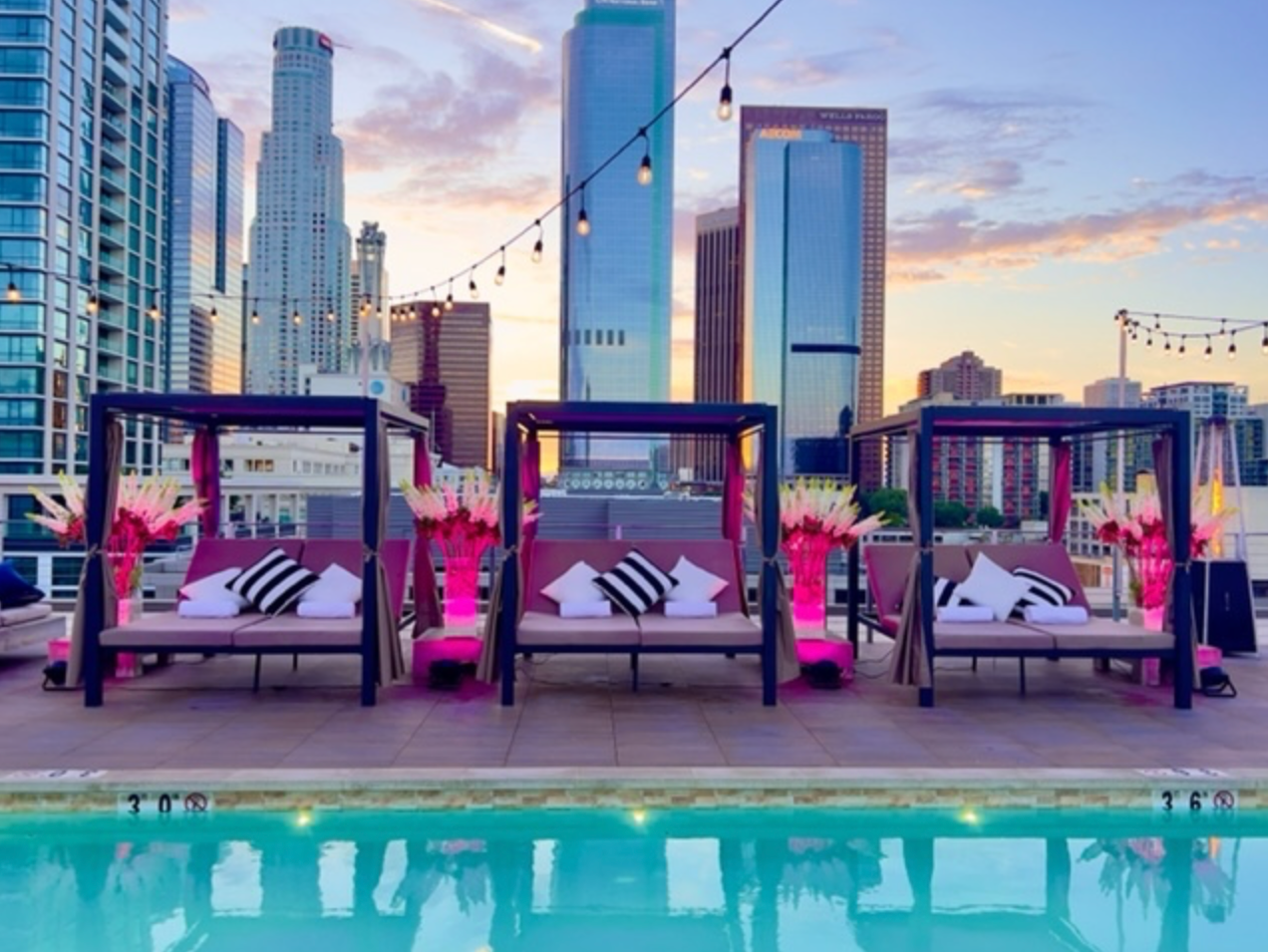 Three cabanas with striped pillows arranged poolside on a rooftop terrace overlooking a city skyline at sunset.