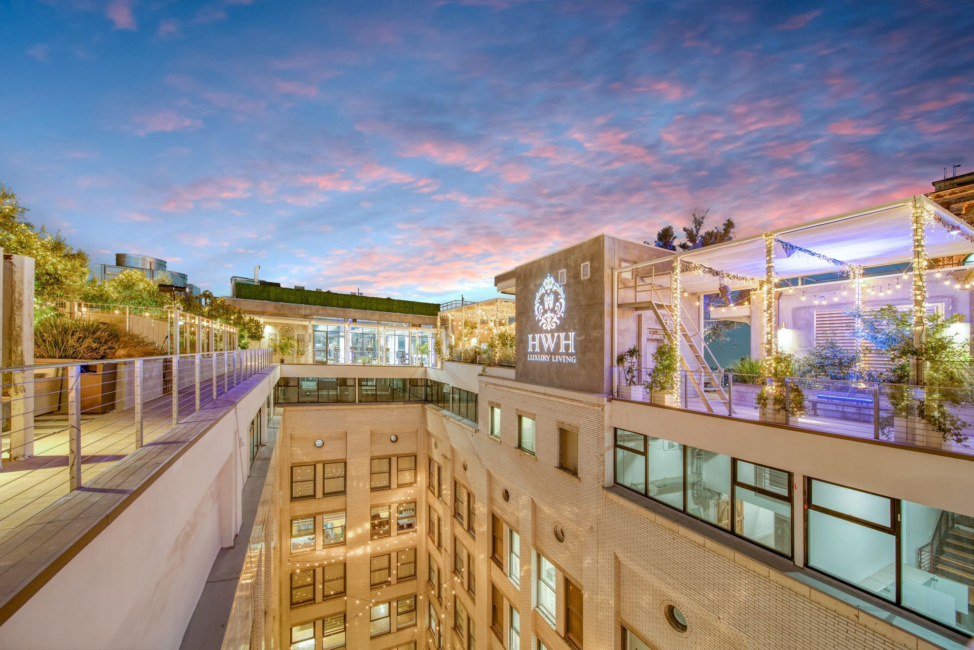 A rooftop restaurant and bar at dusk with a building facade below, illuminated by warm lights under a pink and blue sky.