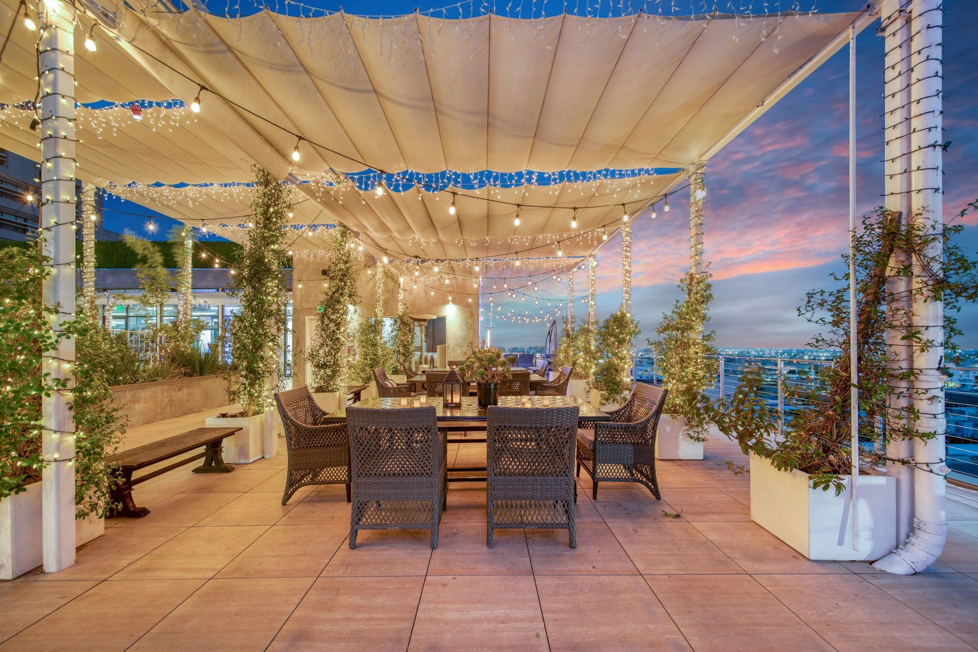 A rooftop patio at sunset featuring a pergola with string lights, wicker dining furniture, and potted plants.