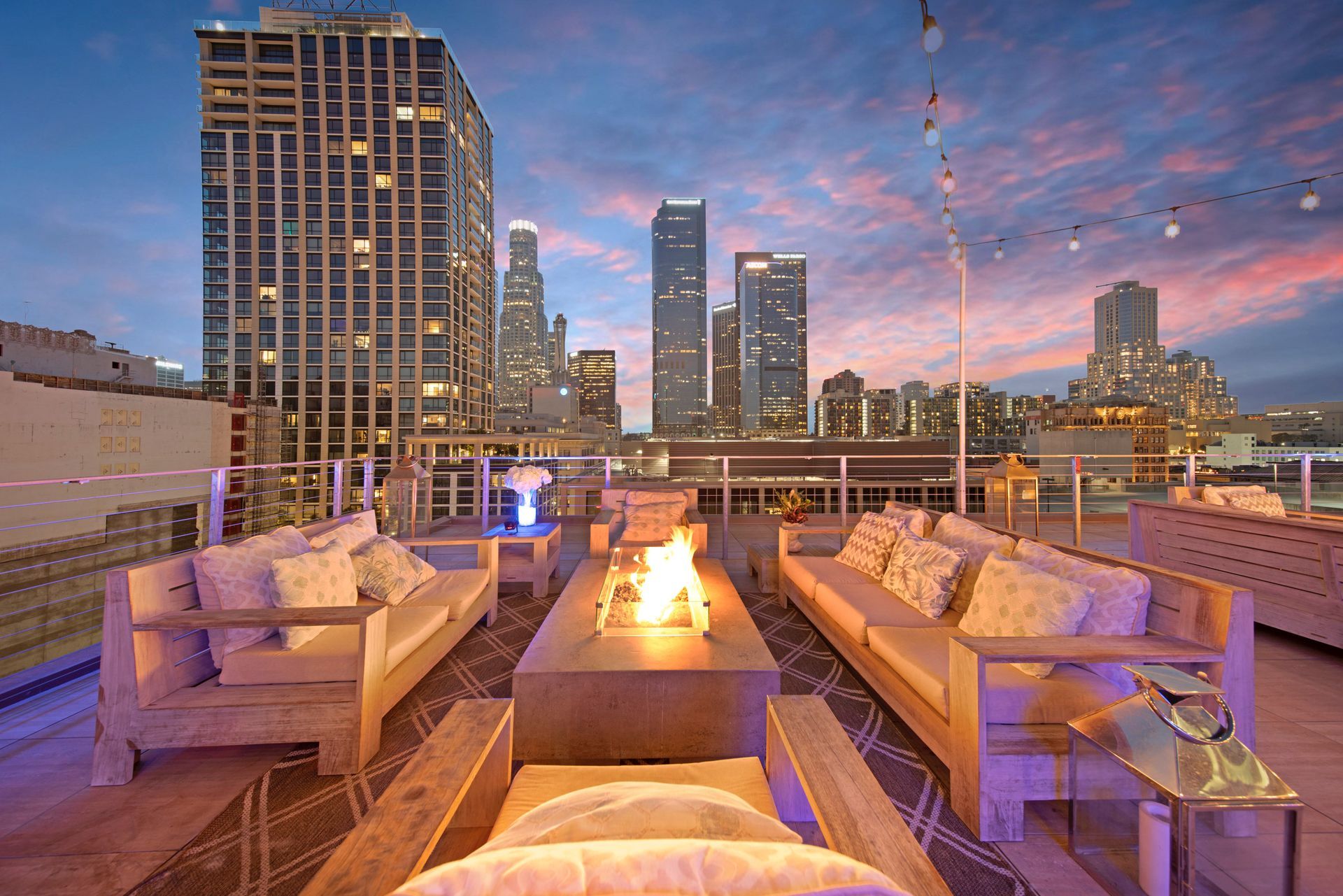 Modern rooftop lounge at sunset with a central fire pit, plush outdoor furniture, and a panoramic view of city skyscrapers.