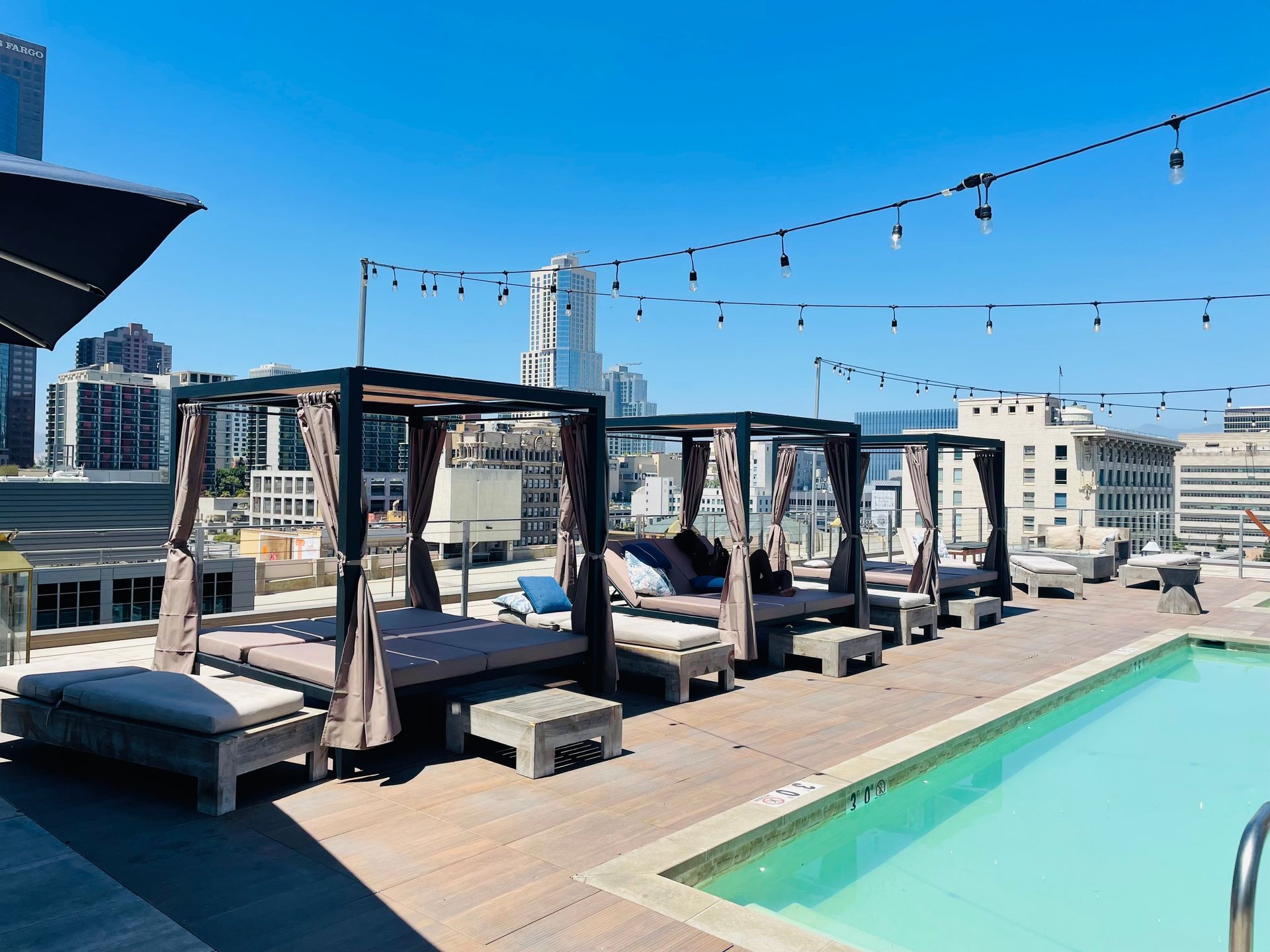 A rooftop pool deck featuring three shaded cabanas with lounge beds alongside a swimming pool under a bright blue sky.
